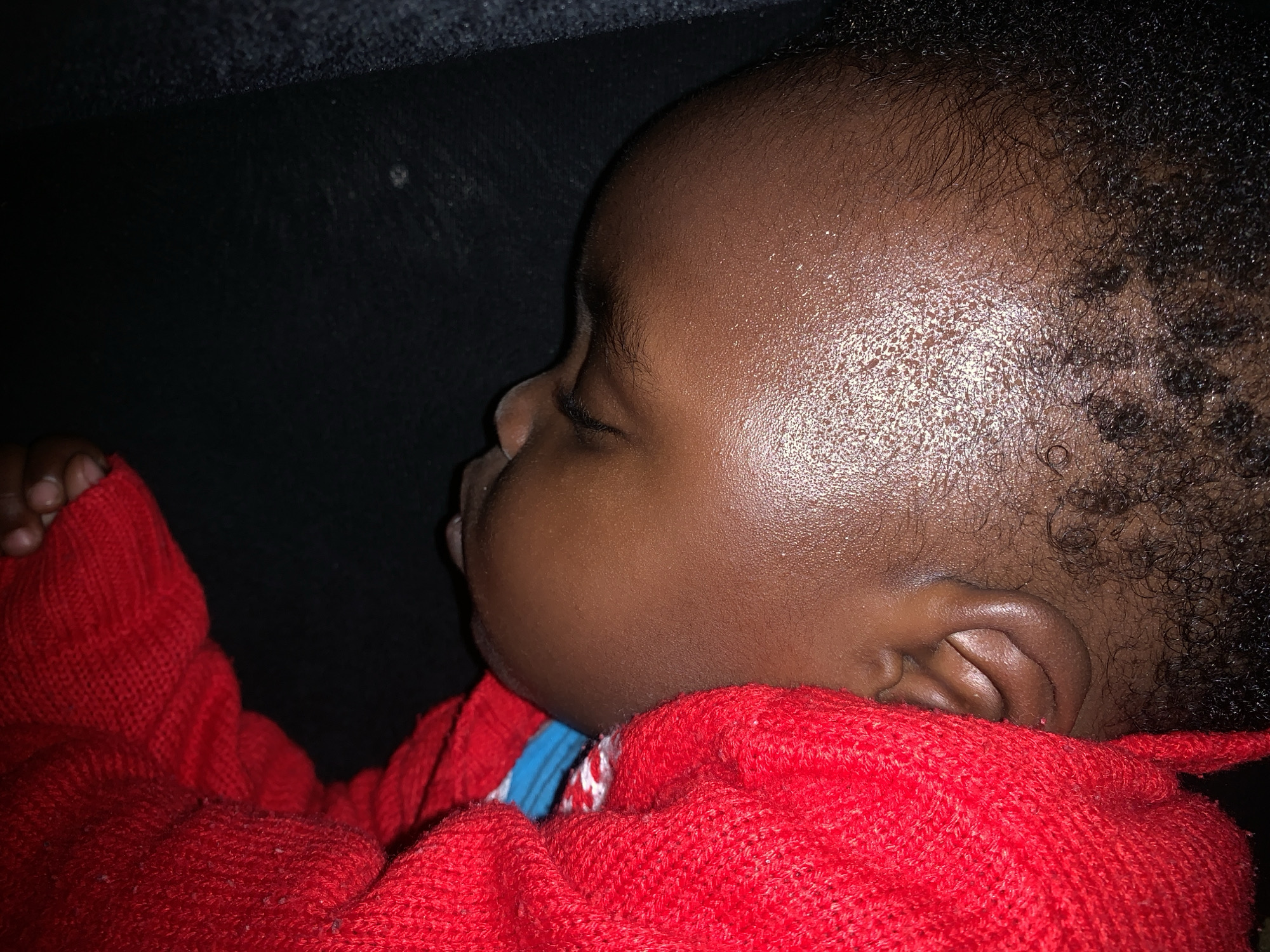 The image shows a close-up, overhead view of a person's head, primarily focusing on their face and ear, which is resting on a dark surface. The person is wearing a bright red knitted garment that covers their shoulder and part of their head. The skin appears to be dark-skinned and is shiny, suggesting perspiration or moisture. The individual's eye is partially visible and closed, indicating they are likely asleep or resting. The hair is dark and appears curly, particularly around the nape of the neck and hairline. The lighting is dim, creating shadows and highlighting the texture of the skin and the knit of the garment. There is no discernible background or environmental context that would suggest a specific location within a city.