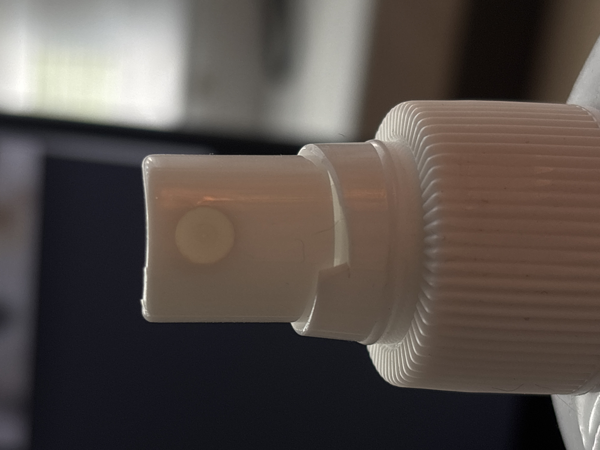 This is a close-up, macro photograph of the nozzle of a white spray bottle, likely containing a liquid product. The focus is on the intricate details of the spray mechanism and the ribbed texture of the bottle's cap. The nozzle itself is a white, slightly rounded piece with a small, circular opening from which the spray would be emitted. A small, circular raised dot or button is visible on the tip of the nozzle. Surrounding the nozzle is a white plastic piece that fits onto the bottle, featuring a clean, smooth finish. The larger part of the bottle cap is visible to the right, characterized by its deeply ridged, ribbed texture, designed for grip and easy twisting. The material appears to be plastic. The background is blurred, with dark blues and grays dominating, suggesting an indoor setting, possibly with soft lighting from a window or overhead light source, hinted at by the bright, diffused white area in the upper portion of the image. There is no visible text. The overall impression is one of a product detail shot, highlighting the design and functionality of the spray bottle's dispensing end. The lighting and shallow depth of field contribute to a clean and modern aesthetic. The location context of Saint Petersburg, Russia, does not provide any specific visual cues within this close-up image of a common object.