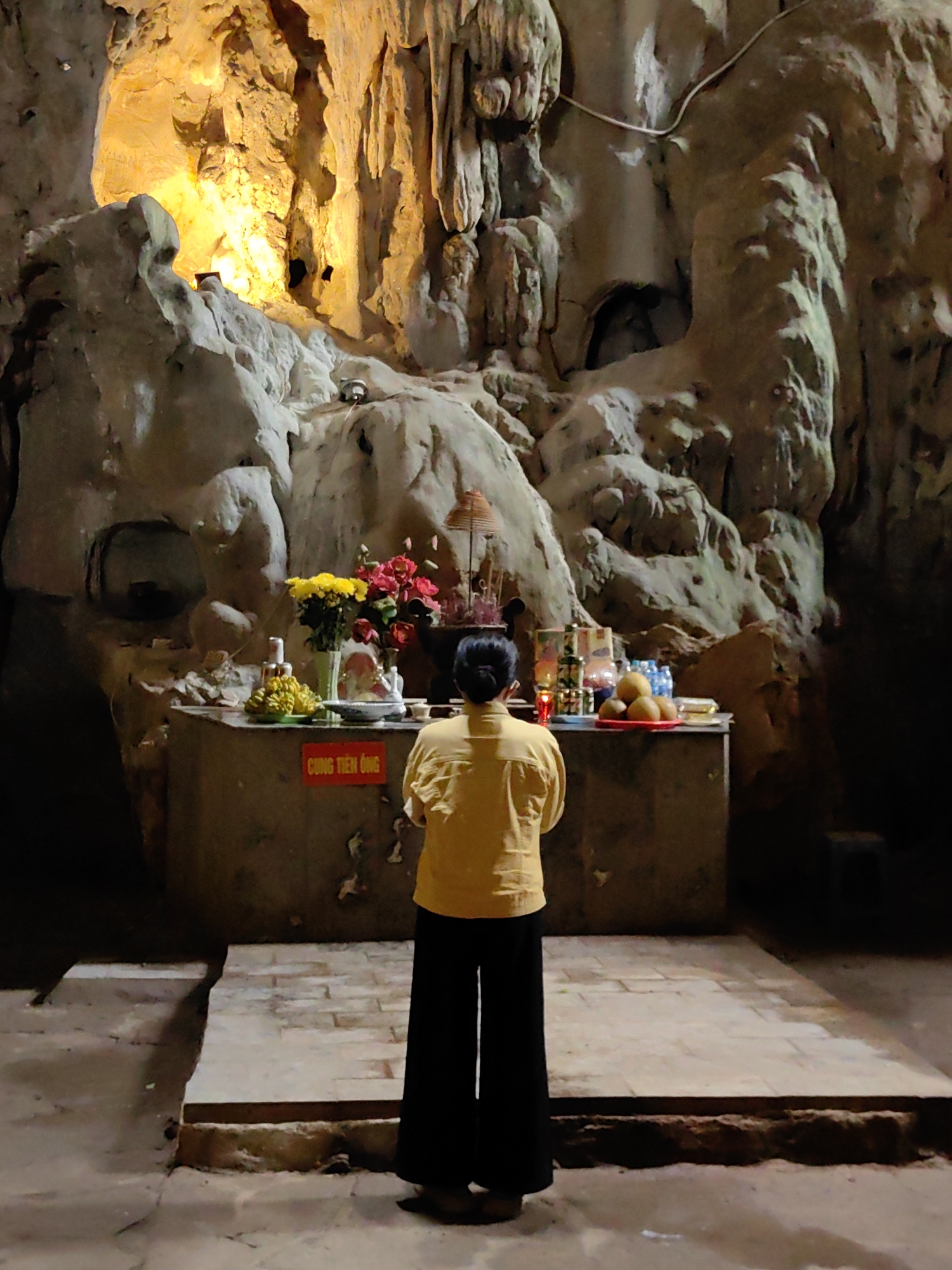 A woman stands facing an altar in a cave, presumably in Lạng Sơn, Vietnam. She has her back to the viewer, wearing a yellow shirt and black pants, with her hands clasped in front of her. The altar is elevated on a stone platform and holds offerings including flowers, fruit, and burning incense sticks. A red sign with yellow text is visible on the front of the altar. The cave's rock formations are illuminated by a light source behind and above the altar, highlighting the textures of the stone. The floor around the altar is rough and uneven.