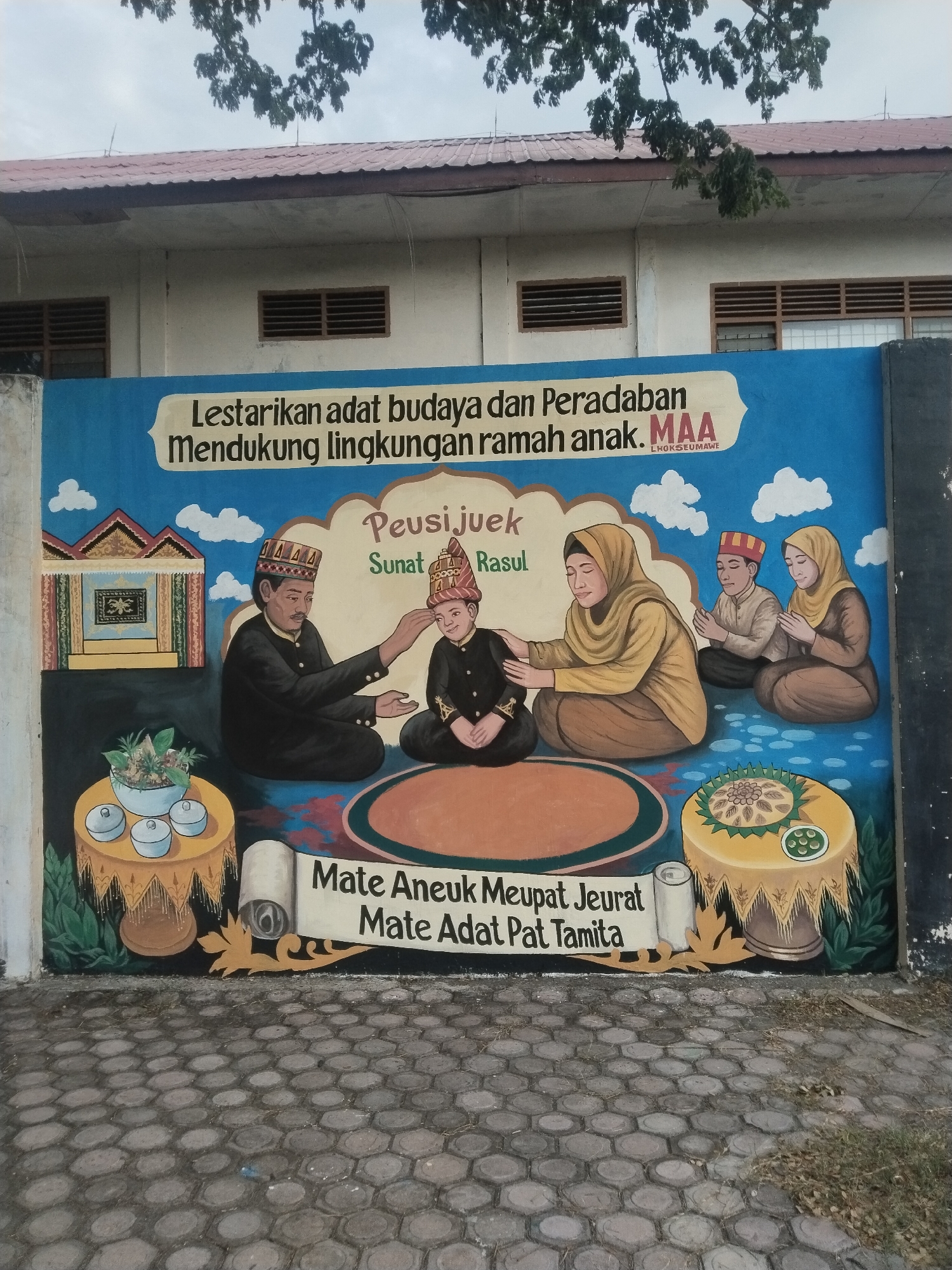A vibrant mural is painted on an outdoor wall in Lhokseumawe, Indonesia, depicting a traditional ceremony. The scene features five individuals seated on the ground. In the center, a young boy in ceremonial attire and a traditional head covering receives a blessing from an adult man, who also wears traditional clothing and a headpiece, placing a hand on the boy's head. An adult woman, wearing a hijab and traditional dress, sits to the boy's right, offering support with a hand on his back. Two other children, a boy and a girl, are seated further to the right, observing with hands clasped in front, dressed in similar traditional attire.
To the left of the main figures, a traditional Acehnese house is depicted. Two ornate, low tables flank the central group: one on the left holds a bowl of flowers or herbs and four small lidded dishes, while the one on the right displays a large, elaborate dish of food and a smaller bowl. The background of the mural is a bright blue sky with white clouds, fading into a darker foreground where the figures are seated.
The text visible on the mural conveys several messages. At the top, a banner reads: "Lestarikan adat budaya dan Peradaban / Mendukung lingkungan ramah anak. MAA / LHOKSEUMAWE" (Preserve culture and civilization / Supporting child-friendly environment. MAA / Lhokseumawe). A central banner highlights the event: "Peusi juek / Sunat Rasul," referring to a traditional Acehnese pre-circumcision ritual and the Islamic practice of circumcision. At the bottom, another banner displays an Acehnese proverb: "Mate Aneuk Meupat Jeurat / Mate Adat Pat Tamita" (If a child dies, its grave is known / If tradition dies, where can it be found?).
The photograph, taken in daytime under an overcast sky, shows parts of a building with windows and a roof above the mural, and a paved hexagonal-block ground in front of the wall. The mural's artwork is illustrative, employing warm colors and clear outlines to portray a significant cultural event with reverence and community spirit.