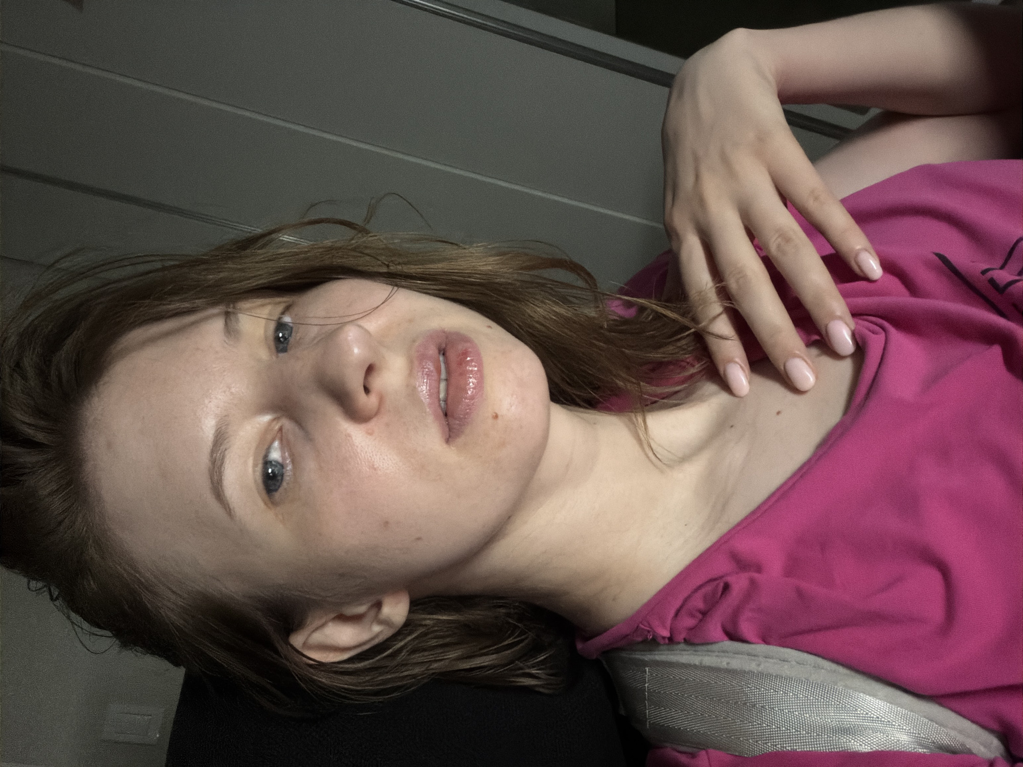 A young Caucasian woman with light brown hair and blue eyes is taking a selfie in a dimly lit indoor setting. She is wearing a bright pink t-shirt and has her left hand resting on her chest. Her lips are glossed and slightly parted. The background appears to be a wall with vertical paneling. The image is a close-up shot, focusing on the woman's face and upper body. There are no other people or objects visible in the frame. The lighting is low, with a light source coming from the front, casting shadows on the right side of her face and body.