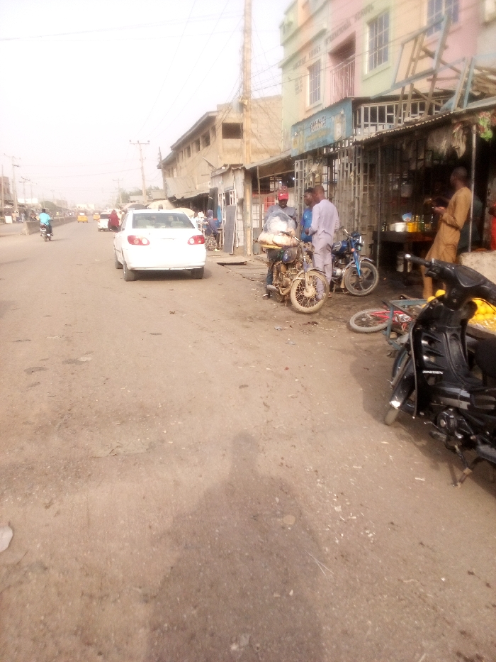 A bustling street scene unfolds with a white car parked on the left, obstructing traffic. Adjacent to a row of buildings with corrugated metal awnings, several individuals gather near motorcycles. One person in traditional attire stands at a makeshift food stall. The road is worn and dusty, revealing the shadow of the photographer. Overhead, power lines stretch across the scene under a muted sky.