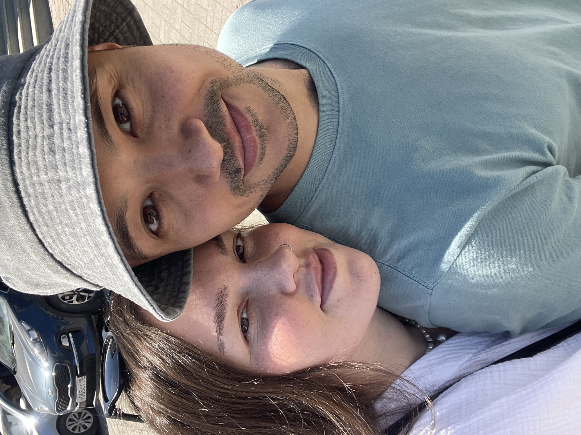 Two individuals, a male and a female, are lying head-to-head, facing upwards towards the camera. The male is wearing a light blue t-shirt and a gray bucket hat. He has brown eyes and a short beard. The female has brown hair and a nose ring. She is wearing a white top. The background features a black car and a paved surface, suggesting an outdoor location. The license plate on the car reads "Y065 CT 18," which indicates the location as Izhevsk, Russia.