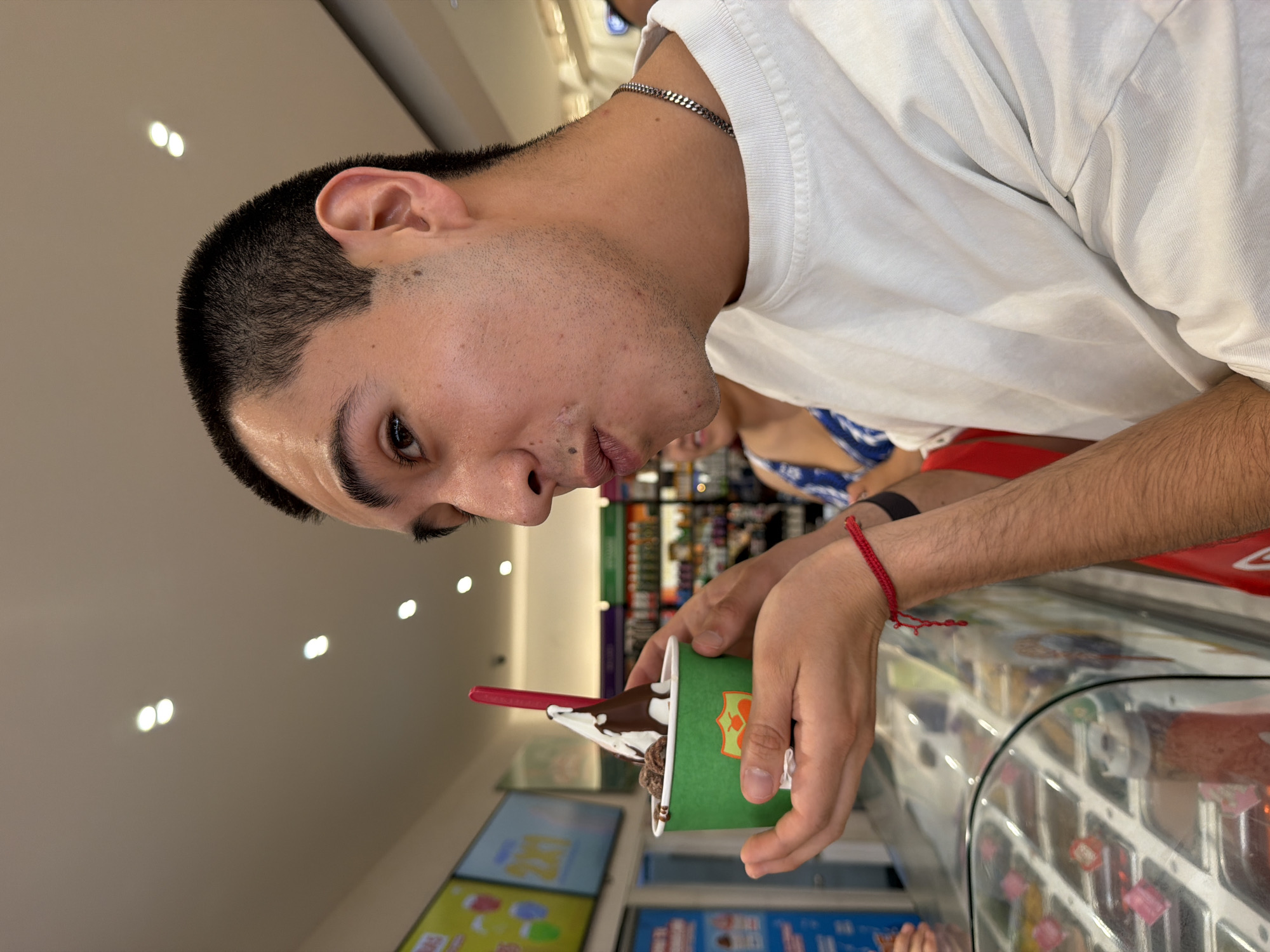 An eye-level indoor shot captures a young man in a frozen yogurt or ice cream shop in San Francisco Ocotlán, Mexico. The subject, a male with short dark hair, is seen from the chest up, looking downwards at a green paper cup he holds with both hands. He is wearing a white t-shirt, a thin silver chain, and a red string bracelet on his right wrist. His lips are slightly pursed as he gazes at the dessert.
The green cup contains a white soft-serve substance topped with dark chocolate sauce and other dark, granular toppings, possibly cookie crumbles. A red spoon is inserted into the dessert. The cup features a partial orange or yellow logo.
The background shows a modern retail environment. To the lower right, a clear glass display case with various colorful toppings is visible. To the left, two rectangular screens display bright, colorful graphics, likely advertisements for the shop's offerings, though no specific text is legible. The ceiling is light-colored with multiple recessed lights, indicating a well-lit interior, suggesting it could be daytime or evening. A blurred figure in the background appears to be wearing a blue and white patterned top and red clothing. The overall scene conveys a casual atmosphere of enjoying a sweet treat.