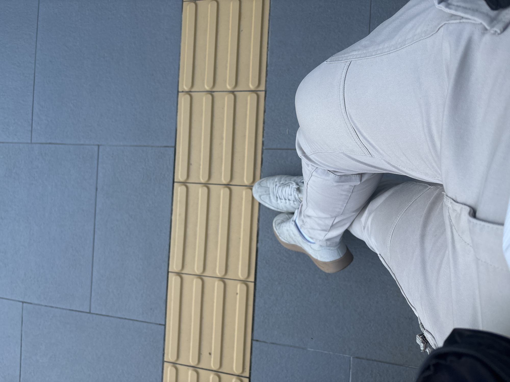 The image is a top-down view of a person wearing light-colored pants and sneakers, sitting on a gray tiled surface next to a tactile paving surface with raised bars. The tactile paving is yellow and is presumably a safety feature for visually impaired pedestrians. The location is in an urban setting, specifically in Sofia, Bulgaria. The person is positioned in relation to the tactile paving, suggesting they are in a public space such as a sidewalk or a train platform. The photograph lacks contextual elements beyond the surface materials and the person's lower body.