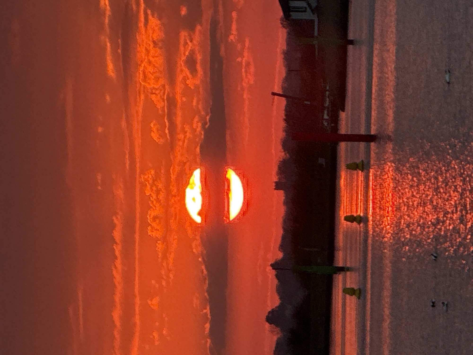 Captured at Great Yarmouth, United Kingdom, the image shows a vivid orange sunset reflecting on water. Two distinct orange-yellow shapes of the setting sun are visible through clouds, with their reflections creating parallel lines on the water's surface. Dock-like structures are silhouetted in the foreground, with small, green objects and vertical poles. The dark silhouette of the land is between the sky and water. Small dark shapes floating on the water could be birds.