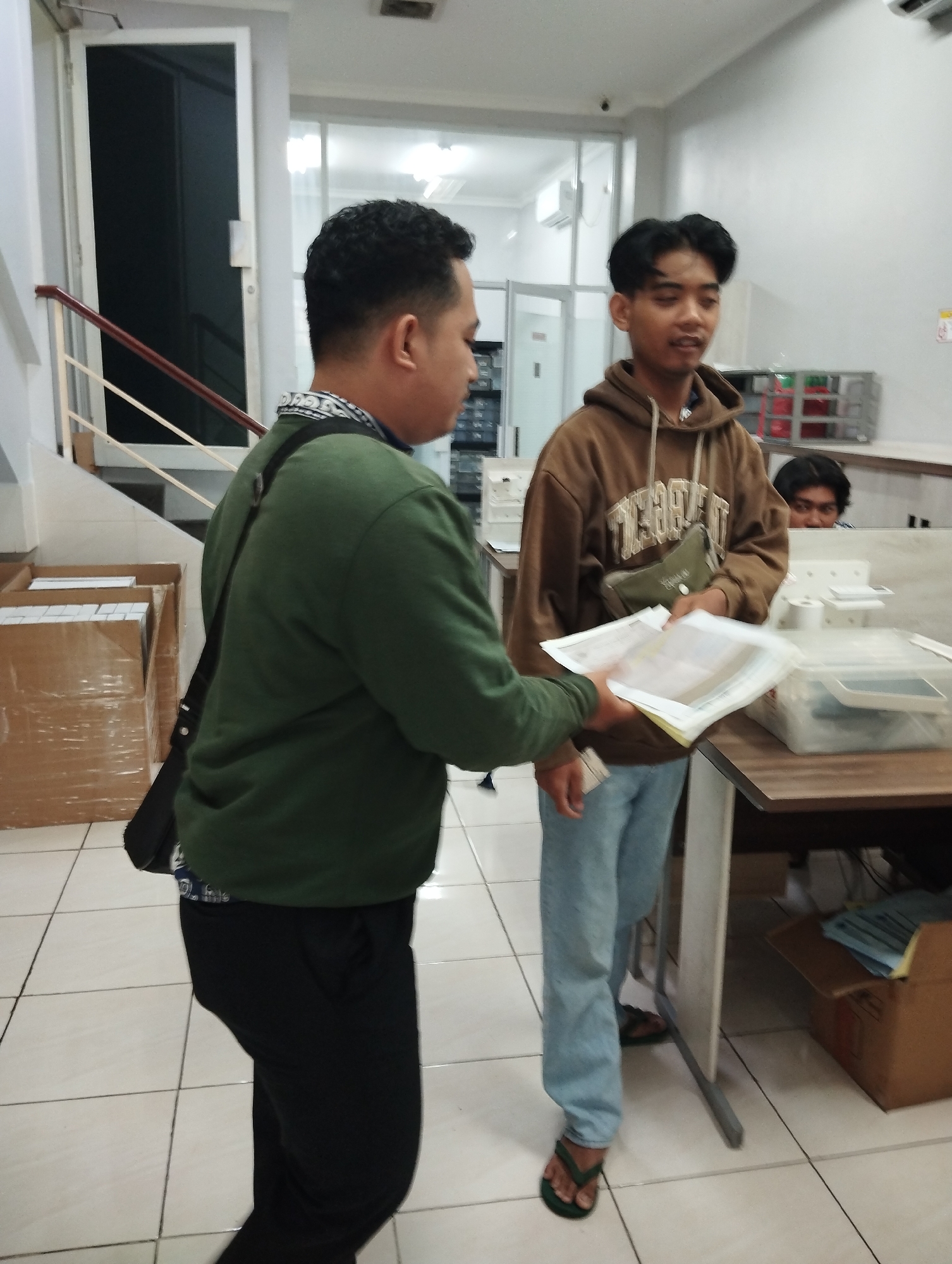 Two young men are interacting in an indoor office environment. The man on the left, wearing a green long-sleeved shirt and black pants, is facing away from the camera and handing a stack of papers to the man on the right. The man on the right, wearing a brown hoodie with a graphic design and light blue jeans, is facing the camera and receiving the papers. He is also wearing a green pouch across his chest. In the background, another person is partially visible behind a partition, looking towards the interaction. The environment appears to be a functional office space with tiled flooring, a desk with office supplies, shelves with items, and a staircase visible in the background. The overall lighting suggests it is daytime or a well-lit interior.