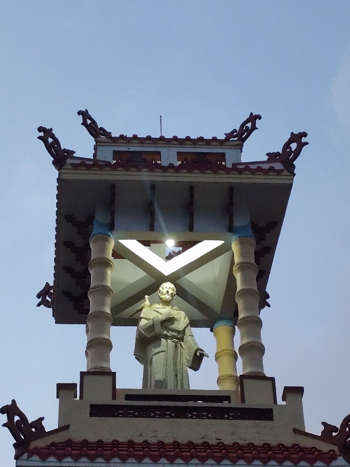 An upward-angled photograph captures the illuminated upper section of a multi-tiered bell tower, set against a light blue-grey sky. At the core of the structure, an elevated, light-colored statue of a bearded male figure, identifiable as Saint Francis of Assisi, stands within an open pavilion. The statue wears a monastic robe, holds an object resembling a book or scroll against its left chest, and has a small bird, possibly a dove, perched on its right shoulder.
The pavilion is supported by four segmented pillars, designed to mimic bamboo stalks, and features a light-colored roof structure with prominent, ornate dark-colored upturned corner decorations. A single bright, circular light fixture is mounted on the underside of the pavilion's roof, directly illuminating the statue. Below this pavilion, a section of a lower tier is visible, characterized by a roof covered in red-brown interlocking tiles and adorned with similar decorative elements at its corners. The walls of this lower tier are light-colored, featuring a dark, horizontally banded decorative frieze. This bell tower is part of the Franciscan Monastery (Tu viện Dòng Phanxicô) in Thành phố Đà Lạt, Vietnam.