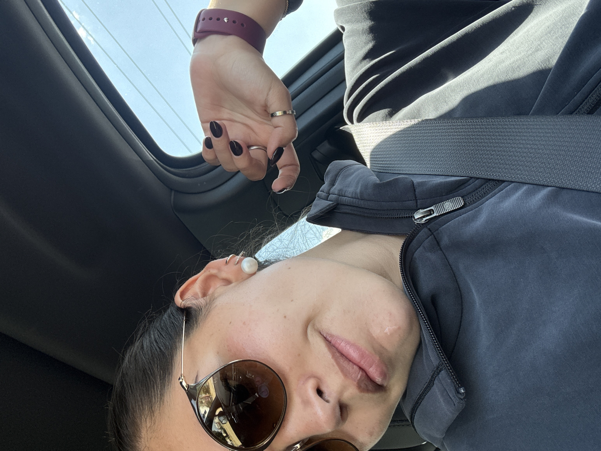 The image captures a partial, angled view of an individual seated in the front of a vehicle during daylight. The subject, an adult, has dark hair pulled back and wears reflective, aviator-style sunglasses. A light-colored mark is present on the chin. The left ear displays a large white pearl stud earring and two small, silver-colored ear cuffs on the helix. The person is wearing a dark grey or black zippered hooded sweatshirt, with a silver-colored zipper pull embossed with a brand logo. A dark grey seatbelt is fastened across the individual's chest and lap. The left hand is raised, revealing dark-colored fingernails, a plain silver-colored band ring on the ring finger, and a burgundy-colored smartwatch on the wrist. Reflections in the sunglasses show outdoor elements, including trees and a distant light-colored vehicle. The background through the vehicle's rear window consists of a clear, bright blue sky with horizontal lines, possibly power lines. The vehicle's interior trim, including the headliner and pillar, is dark-colored. The scene is located in the city of San Bernardino Tlaxcalancingo, Mexico.