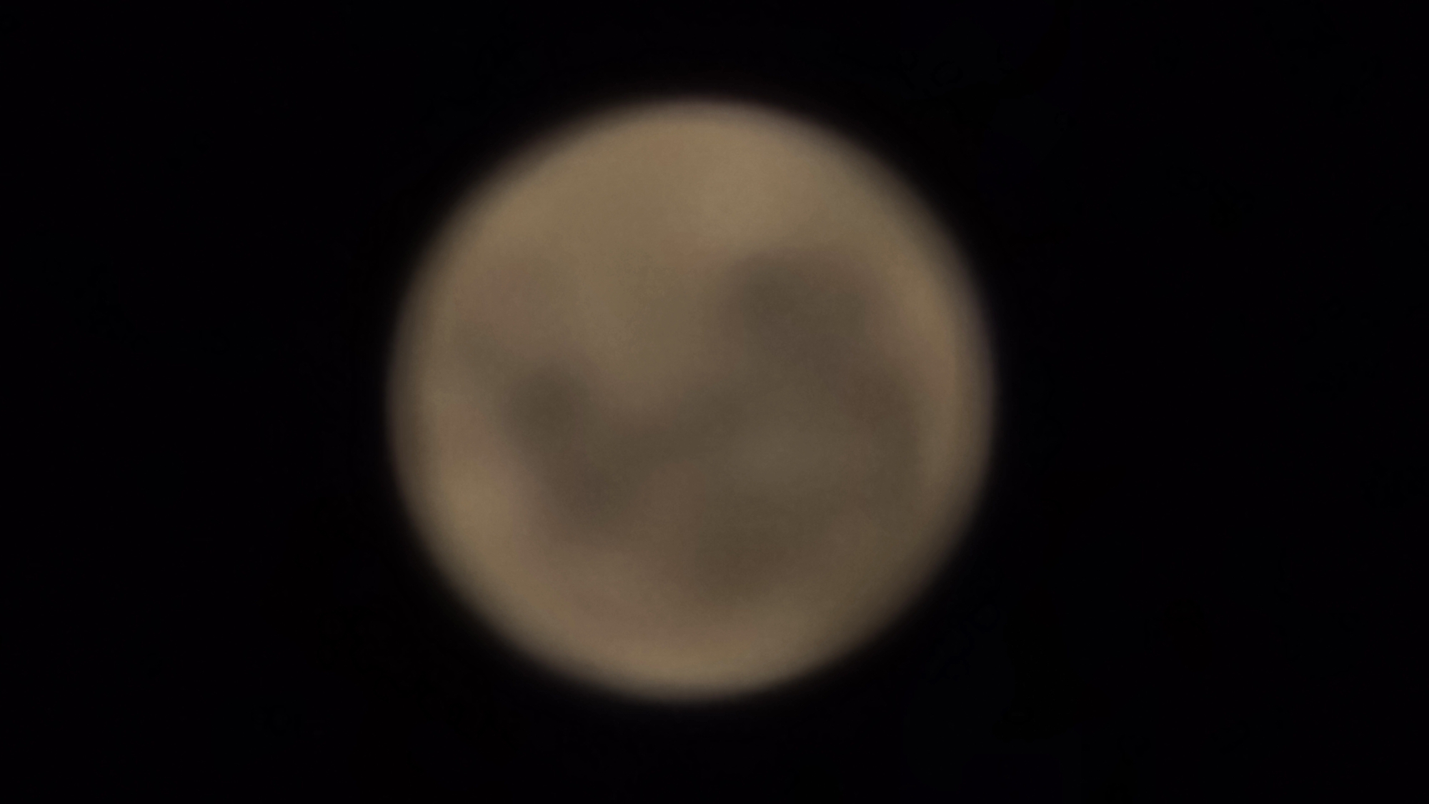 A slightly out-of-focus, circular image of the moon is centered against a black background, suggesting it was taken at night. The moon has a yellowish-brown hue, and faint, darker grey markings are visible on its surface, indicating lunar features such as maria or craters, though their details are obscured by the blur. The overall impression is that of a celestial body observed from Earth. The image does not contain any contextual information that would allow for its localization in Denpasar, Indonesia, or any other specific geographical location.