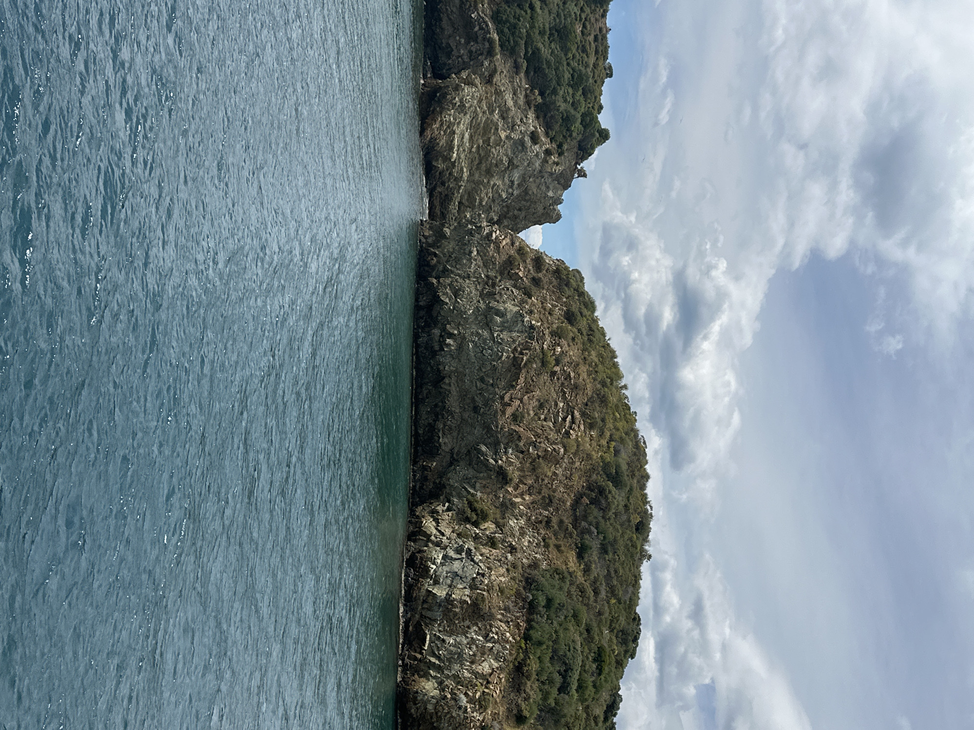 The image depicts a coastal scene, likely in Unknown, Türkiye, featuring a body of water, possibly the sea, occupying the left side of the frame. On the right, two rocky landmasses rise out of the water, sparsely covered with green vegetation. The sky above is filled with a mix of white and gray clouds, suggesting overcast or partly cloudy weather. The water surface shows ripples and subtle reflections of light. No people are visible. The overall composition suggests a natural landscape, possibly a coastal area or a narrow channel between rocky islands.