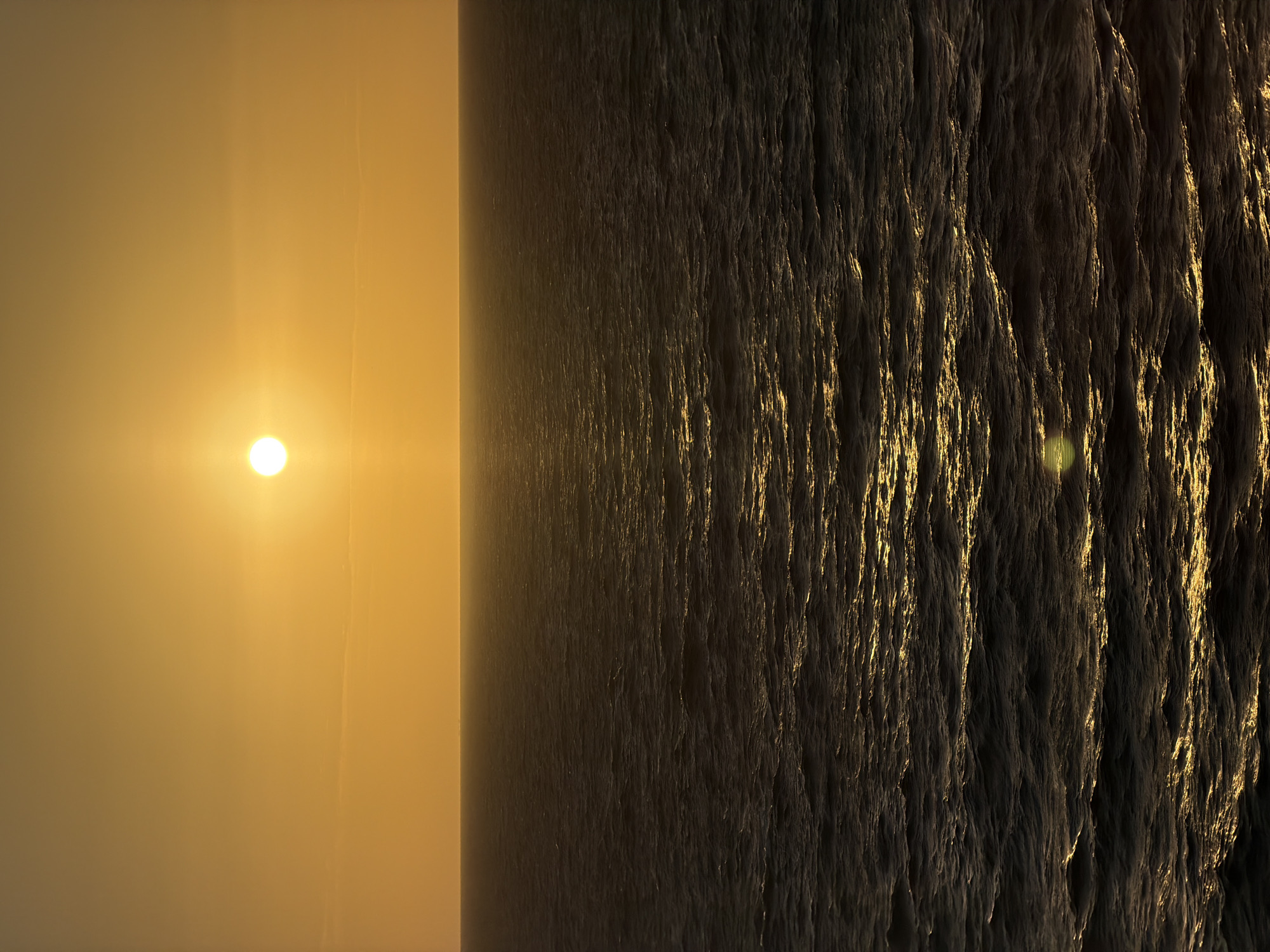 The image depicts a sunset over an ocean or large body of water. The sun is a bright, circular orb low in the sky, casting a golden glow across the water's surface. The water itself appears textured and reflective, with varying shades of dark and light creating a sense of depth and movement. The environment is serene and tranquil, with a clear horizon line separating the sky and water. Location: Unknown, Unknown.