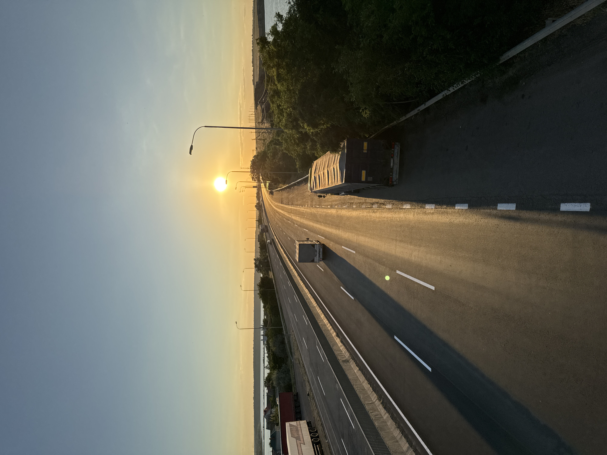 The image depicts a highway at sunset with a clear sky and the sun low on the horizon. The highway is paved and has lane markings. There are several trucks on the highway, some moving and some parked. On the left side of the highway, there are parked trucks and a body of water with some buildings visible in the distance. On the right side of the highway, there is lush green foliage and a body of water. Streetlights line the highway. The overall scene suggests a transportation hub or a scenic route within or near Altestove, Ukraine, characterized by the presence of commercial vehicles and natural landscapes bathed in the warm glow of the setting sun.