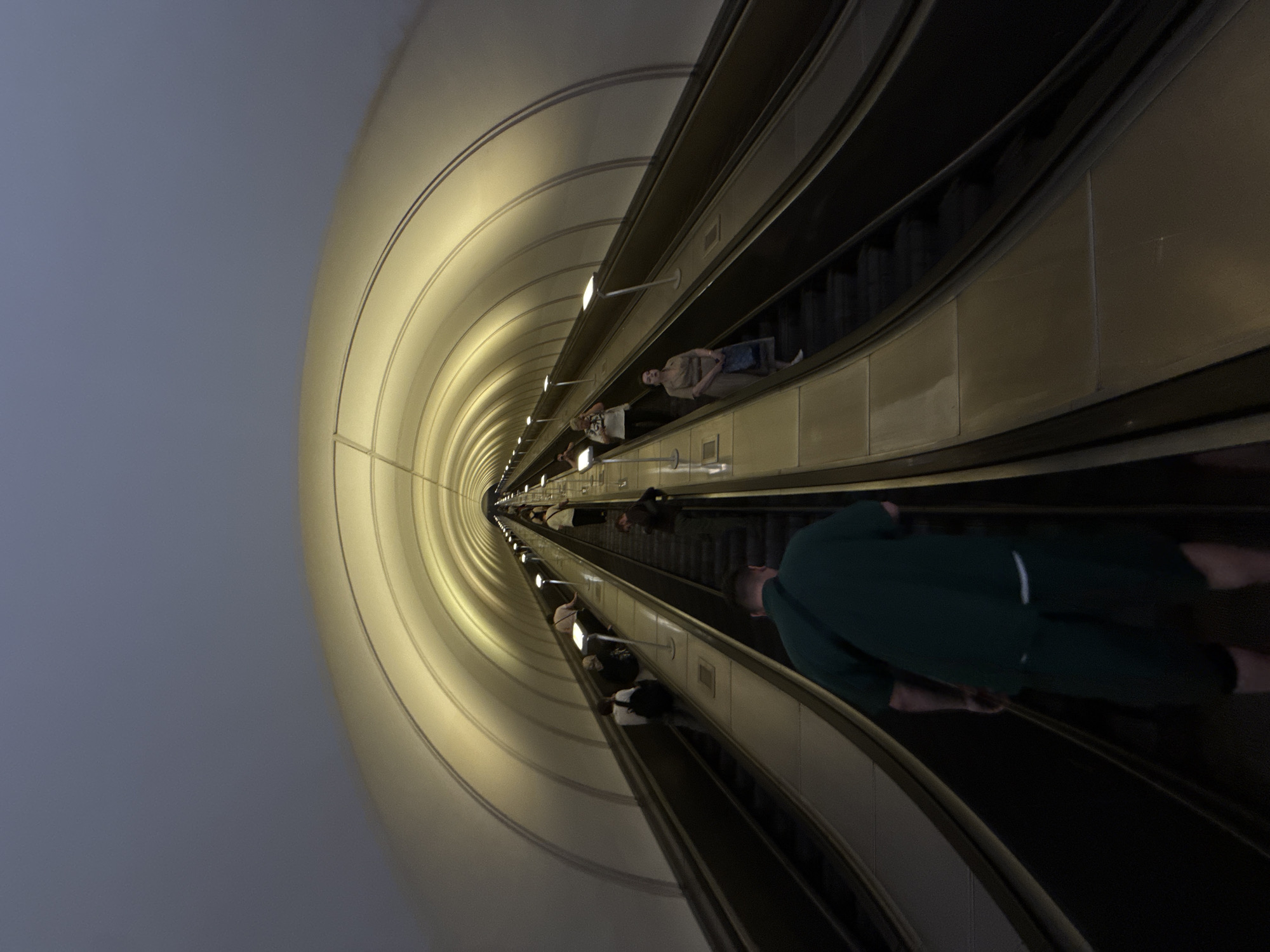 The image depicts a wide, illuminated escalator tunnel in the Moscow Metro, possibly at the VDNKh station due to its distinctive architectural style featuring a curved, ribbed ceiling with recessed lighting. The perspective is from the bottom of the escalator, looking upwards and into the distance. Several people are visible ascending the escalators, which are arranged in parallel banks. In the foreground, a man wearing a dark green t-shirt and shorts is moving up the escalator. Further up, on the right escalator, a woman in a light-colored dress carries a blue bag. On the left escalator, a person with a backpack and another individual are ascending. The tunnel walls are a pale, reflective material, contributing to the overall bright and cavernous feel of the space. The lighting casts long shadows and highlights the repetitive geometric patterns of the tunnel's construction.