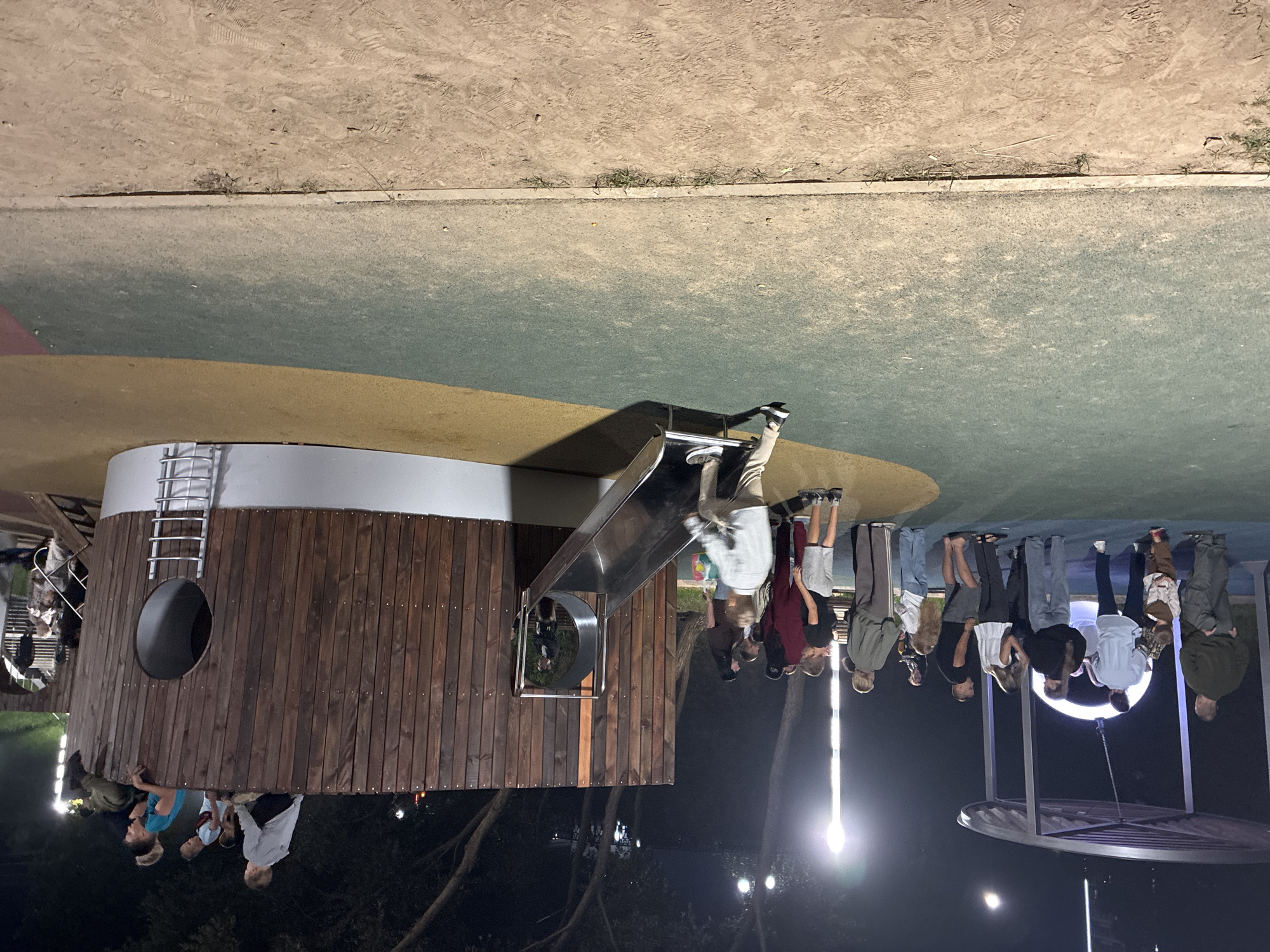 This image depicts an outdoor playground area at night, illuminated by artificial lighting. The playground features a wooden structure resembling a ship, complete with a slide and circular openings. Several people, including adults and children, are present in the scene. Some are gathered near a covered structure with a circular light, while others are near the wooden ship. A young boy is actively going down the slide. The ground is covered with a combination of green and yellow material, transitioning to a dirt path at the bottom of the frame. The background shows trees and more lighting fixtures. The scene is consistent with a public park or recreational space in the city of Mahilyow, Belarus, likely used by families for evening recreation.