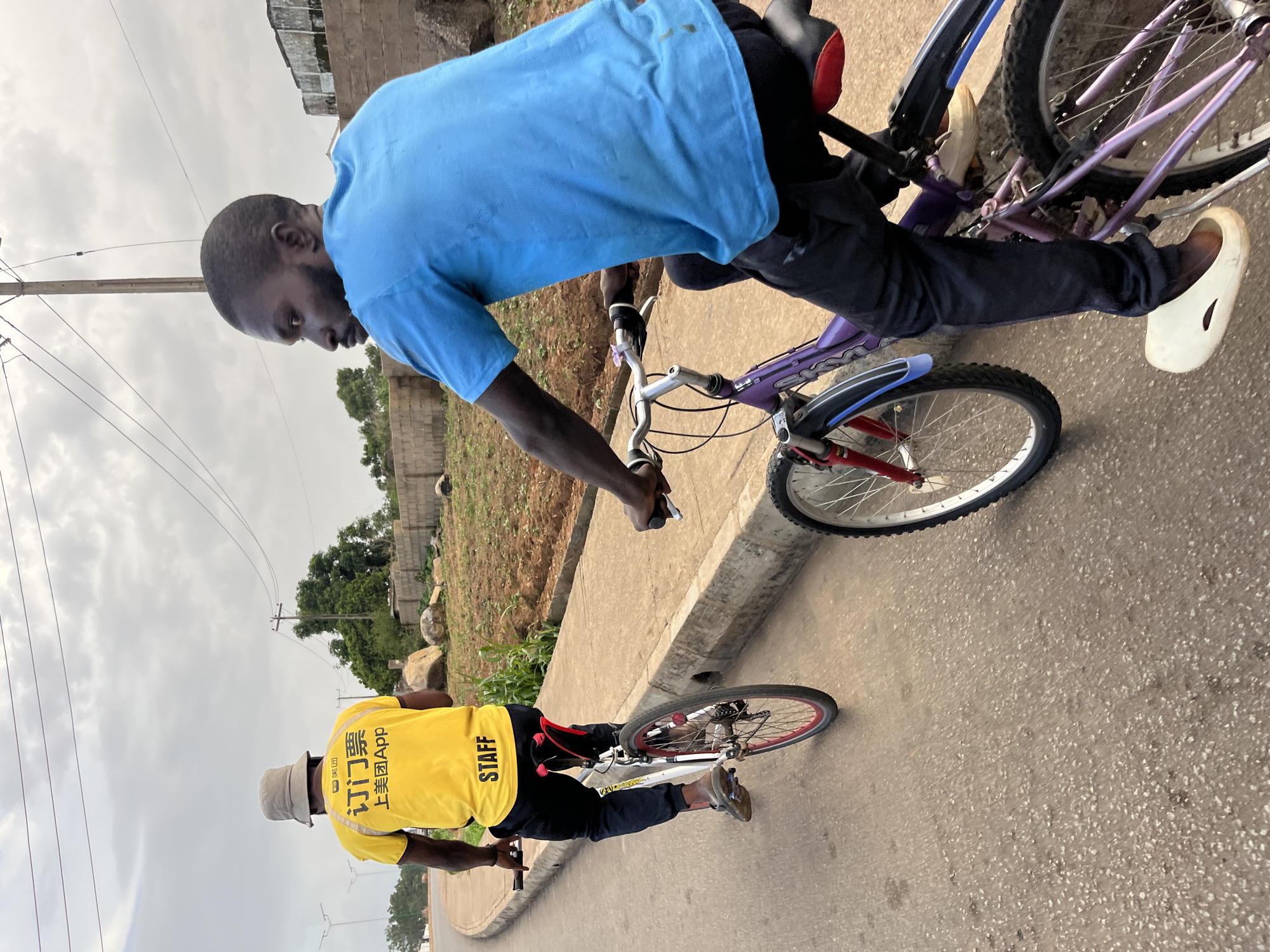 Two individuals are riding bicycles on a paved road in Bukuru, Nigeria. The person in the foreground, on a purple bicycle, is wearing a blue t-shirt and dark pants. They are looking to the left and appear to be in motion. The person in the background, on a white bicycle with red accents, is wearing a yellow t-shirt with Chinese characters and "STAFF" printed in black on the back, along with a light-colored bucket hat and dark pants. This individual is also in motion and facing away from the camera. The environment consists of a paved road, a concrete curb, a patch of dry earth with some greenery, and a stone wall in the background. The sky is overcast with clouds. Electrical poles and wires are visible in the upper portion of the image.