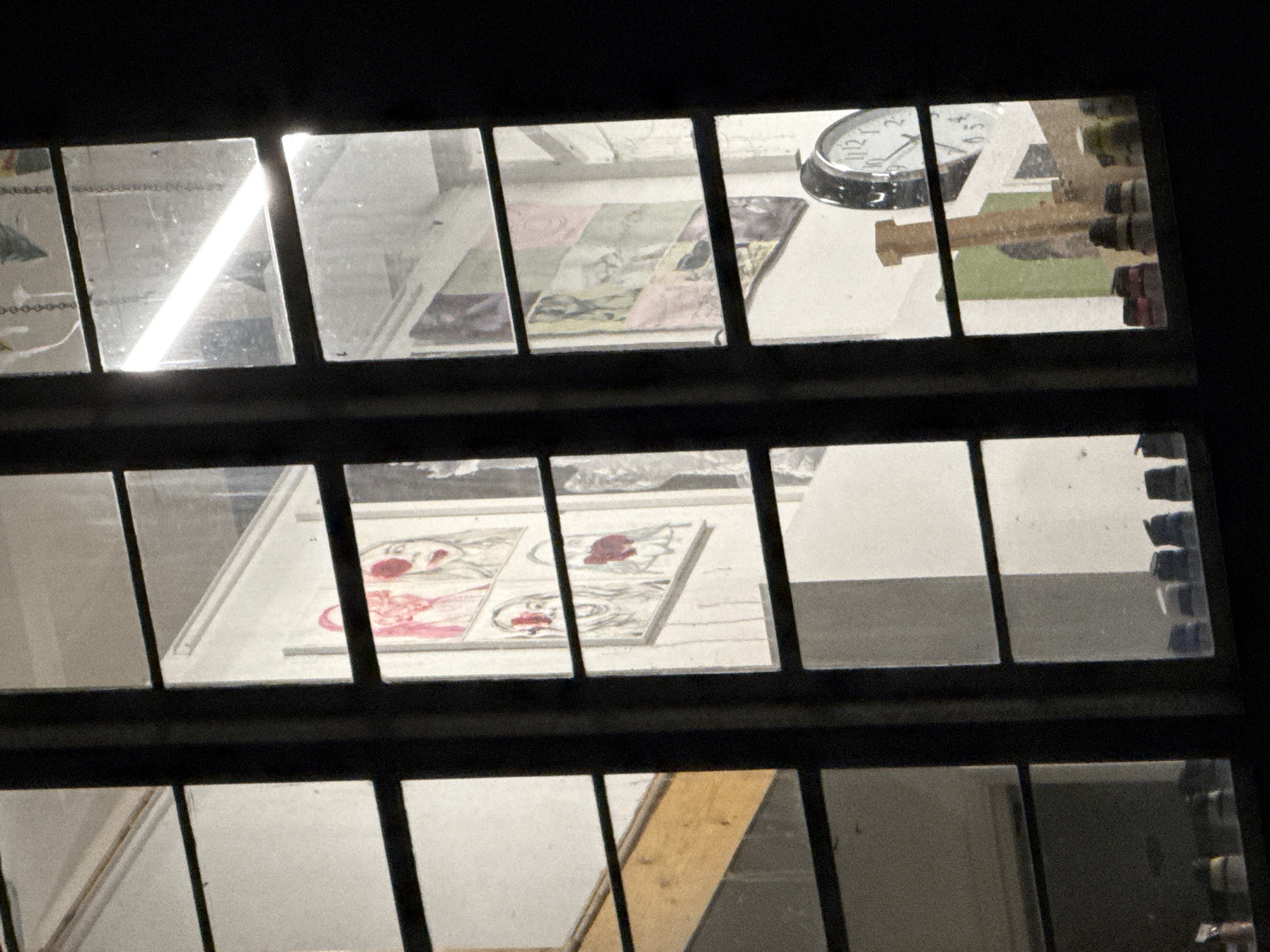The image provides a nocturnal view through a multi-paned window into an illuminated interior, likely an artist's studio or creative workspace in London, United Kingdom. The dark, thick window frames divide the scene into multiple rectangular sections.
Through the upper left panes, a bright, diagonal streak of light from an internal source is visible, reflecting off or shining through the glass. Adjacent to this, a piece of artwork with colorful, patterned segments, resembling a quilt or abstract composition, can be seen. In the upper right section, a silver-rimmed analog clock shows the time around 12 o'clock, with all twelve numbers clearly visible. Near the clock are various tools or supplies, including several brown, cylindrical items and a light green block, indicating a functional workspace.
The middle row of window panes reveals several individual artworks, possibly prints or paintings, framed or matted in white. These pieces predominantly feature stylized faces or figures with striking red elements, such as lips or abstract markings. Below these artworks, the lower section of the window offers glimpses of other structural elements within the room, including a wooden plank angled upwards and other indistinct surfaces like walls or shelving.
No people are visible. The scene captures a static moment, suggesting a late-night setting for artistic production or display. The focus is on the materials and output of a creative endeavor, viewed from the outside or an adjacent dark space.