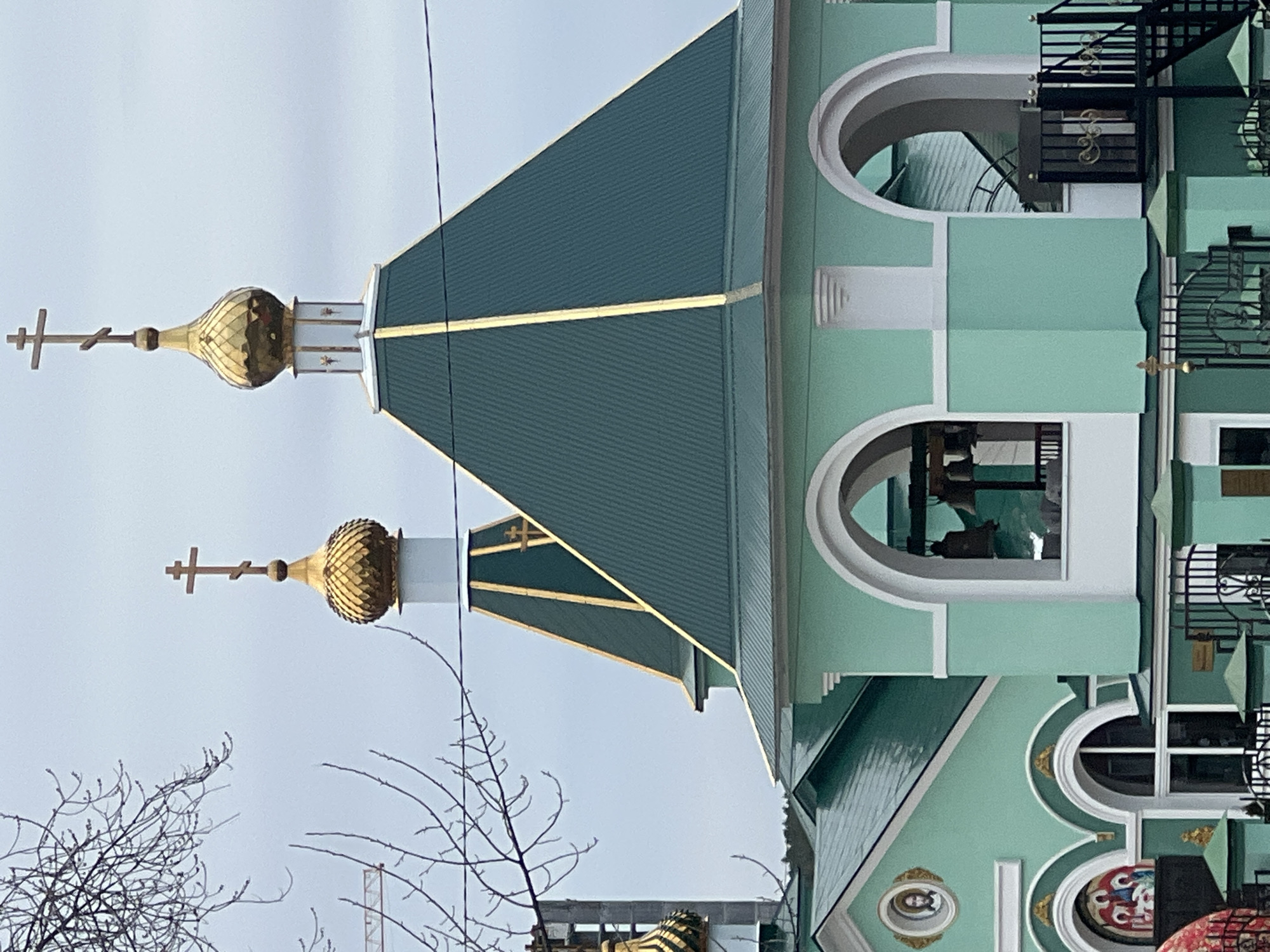 The image shows a close-up of the Church of St. Nicholas, located in Chaikovsky, Russia. The architectural details include green and white painted exterior, golden domes with Orthodox crosses atop white cylindrical structures. The roof is dark green with golden trim. The building features arched windows and a black iron fire escape. Bare branches are visible in the foreground against a pale sky.