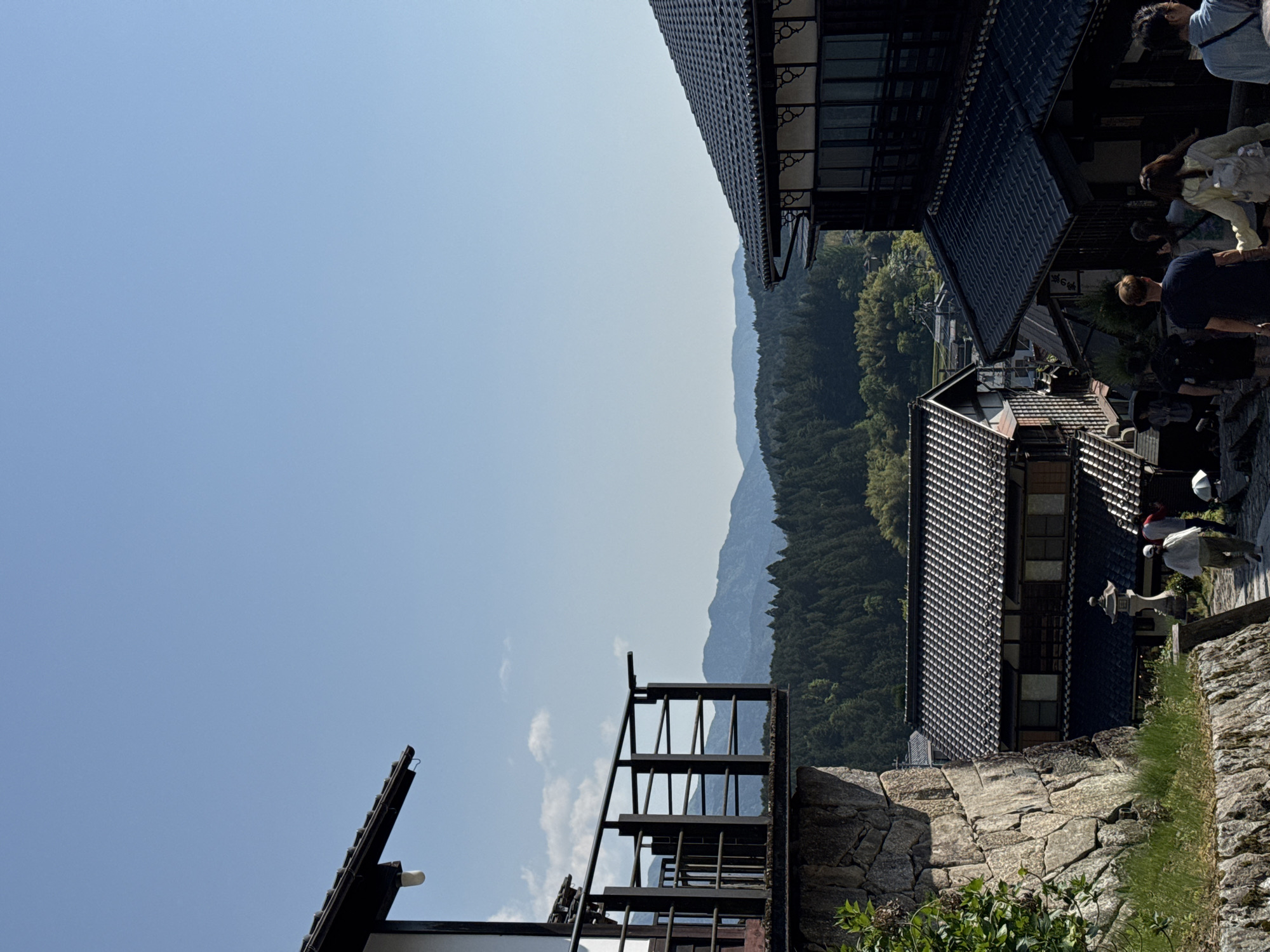 The image depicts a partial view of a street scene in the city of Nakatsugawa, Japan, characterized by traditional Japanese architecture.
Several buildings are visible, featuring dark tiled roofs and wooden frameworks. A stone wall is in the foreground and structures include a set of metal stairs, roofs, and architectural elements.
In the background, forested mountains are visible.
Several people are present in the upper right portion of the image.