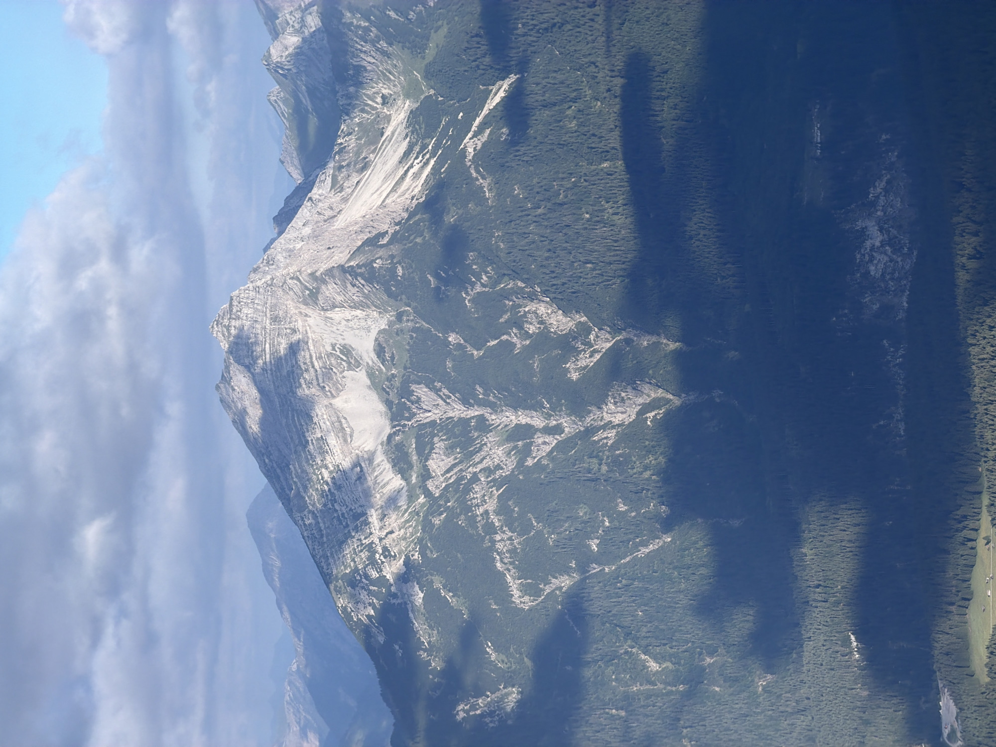 The image shows a panoramic view of the Alps mountain range. In the foreground, a dense, dark green forest covers the lower slopes of the mountains. Shadows from clouds cast large, dark patches across the forest. Higher up, the mountains become rocky and bare, with visible layers and striations in the rock face. A prominent, jagged peak dominates the center of the frame, its summit catching the sunlight. The sky above is a mix of blue and scattered, wispy clouds. The scene appears to be a natural landscape, with no visible human structures or activity in the immediate vicinity of the mountains themselves, though a few small buildings and cleared patches of land are visible in the far distance at the very bottom of the image, suggesting the presence of a village or settlement. Based on the context provided, this scene depicts a view of the Alps near Grainau, Germany, likely taken from a vantage point that overlooks the mountain range.
