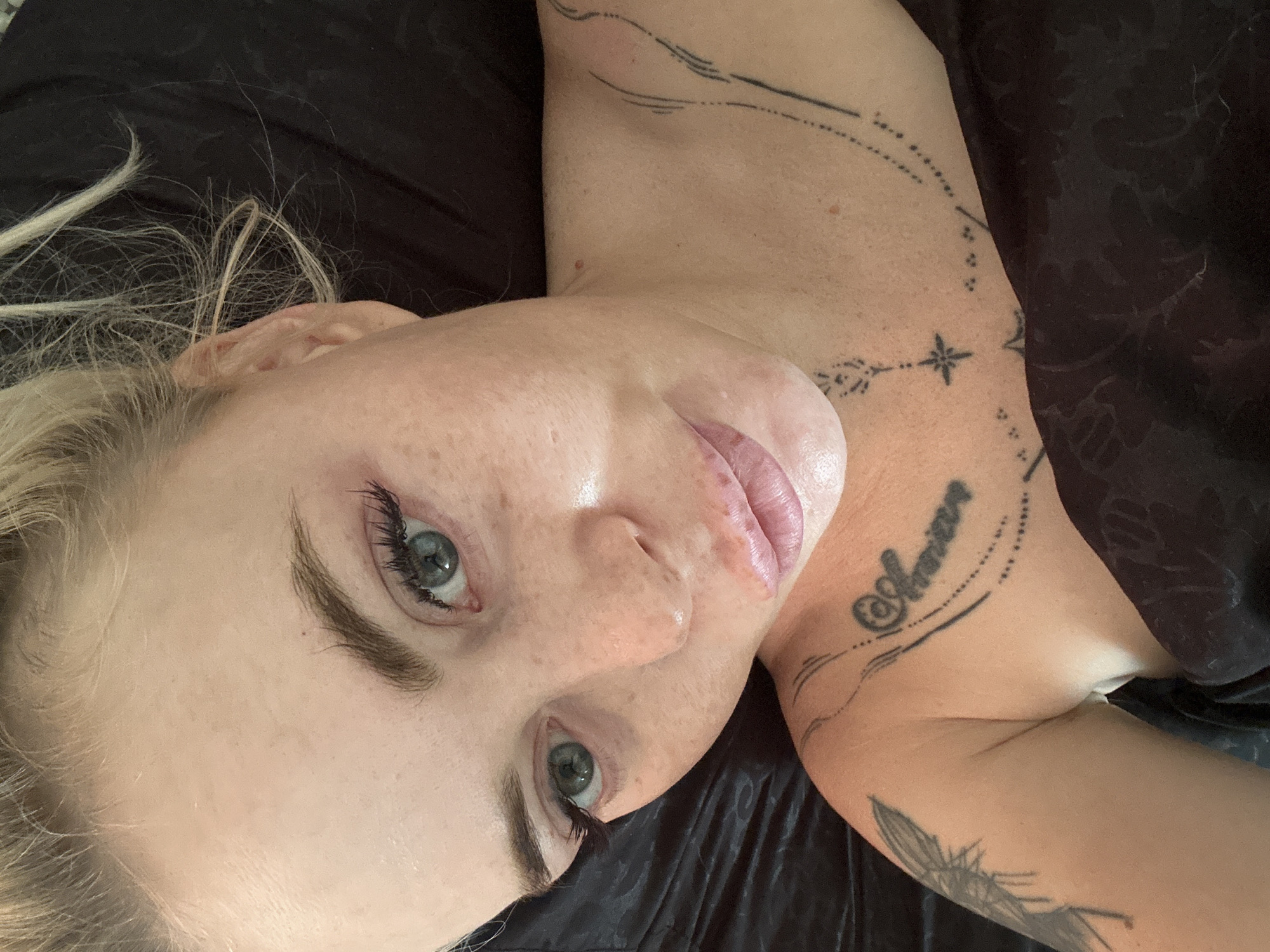 The image is a close-up, high-angle selfie of a blonde woman with light-colored eyes and freckles, lying down. She is wearing makeup, including mascara and lipstick, and has false eyelashes. Her hair is pulled back loosely. Visible on her chest and shoulder are several tattoos: a script tattoo on her collarbone, a star-like design on her chest, a dotted line design extending from her collarbone, and a winged creature on her upper arm. The background is dark and appears to be fabric, possibly bedding. The woman's face is illuminated from above, casting soft shadows.