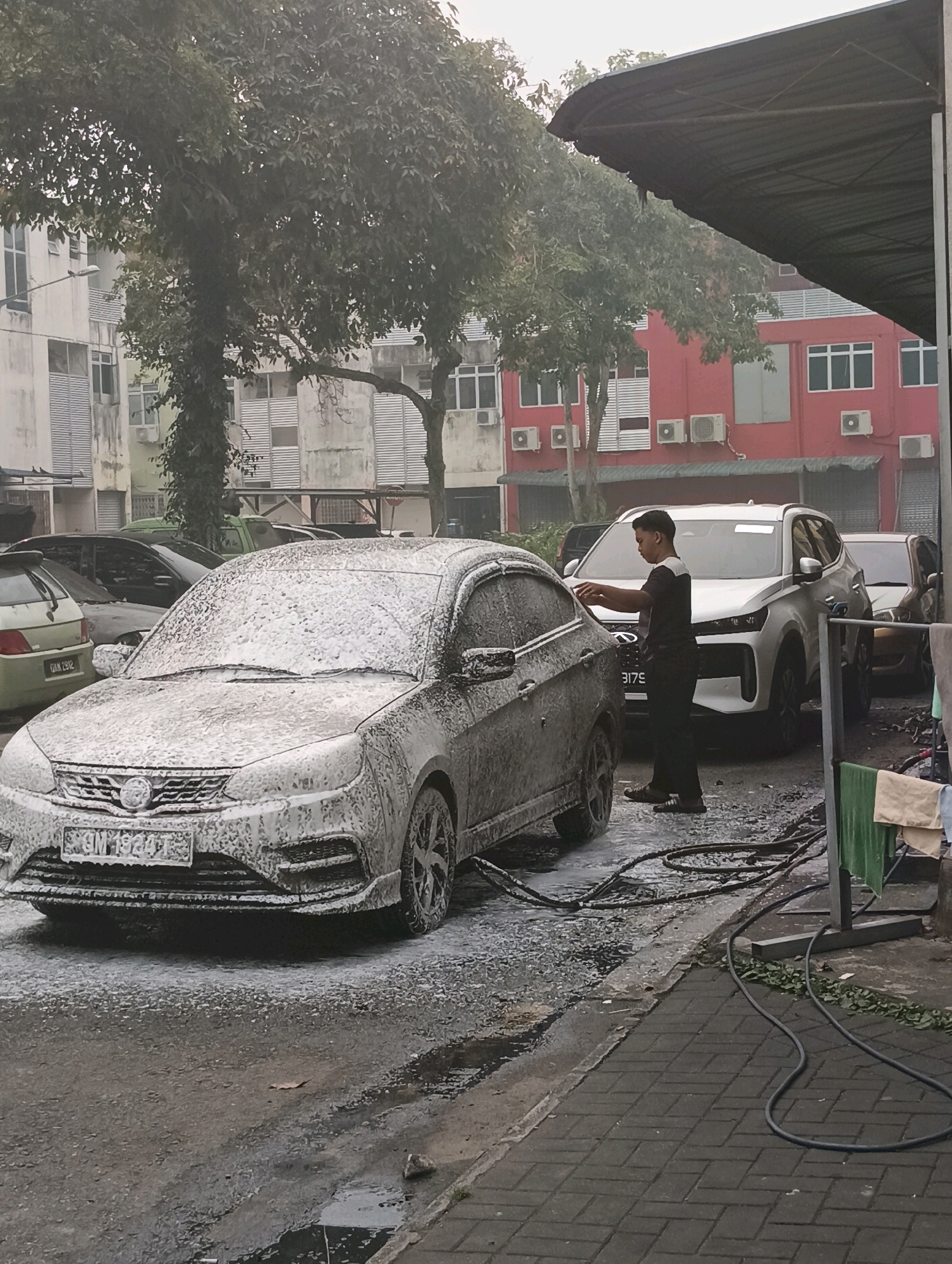 A young man is washing cars in an outdoor car wash bay in Kuching, Malaysia. The scene is set against a backdrop of apartment buildings. The foreground features a silver car heavily covered in white soap suds. To the right of this car, a modern white SUV is partially visible, and a young man wearing a dark t-shirt and dark pants is actively engaged in washing the car, holding a spray nozzle. The ground is wet and soapy, indicating recent or ongoing washing activity. Several other cars are parked in the background. The environment appears to be overcast, with a large dark canopy providing partial shelter over the car wash area. The overall impression is a typical urban car wash service.