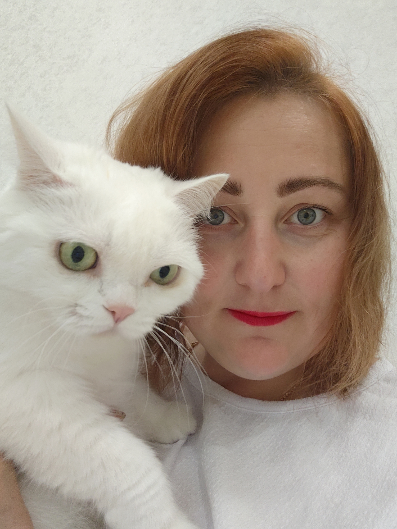 A white cat with green eyes is being held by a woman with red hair and blue eyes. The woman is wearing a white shirt and red lipstick. The background is a plain white wall. The image was taken in the city of Markovskii, Russia.