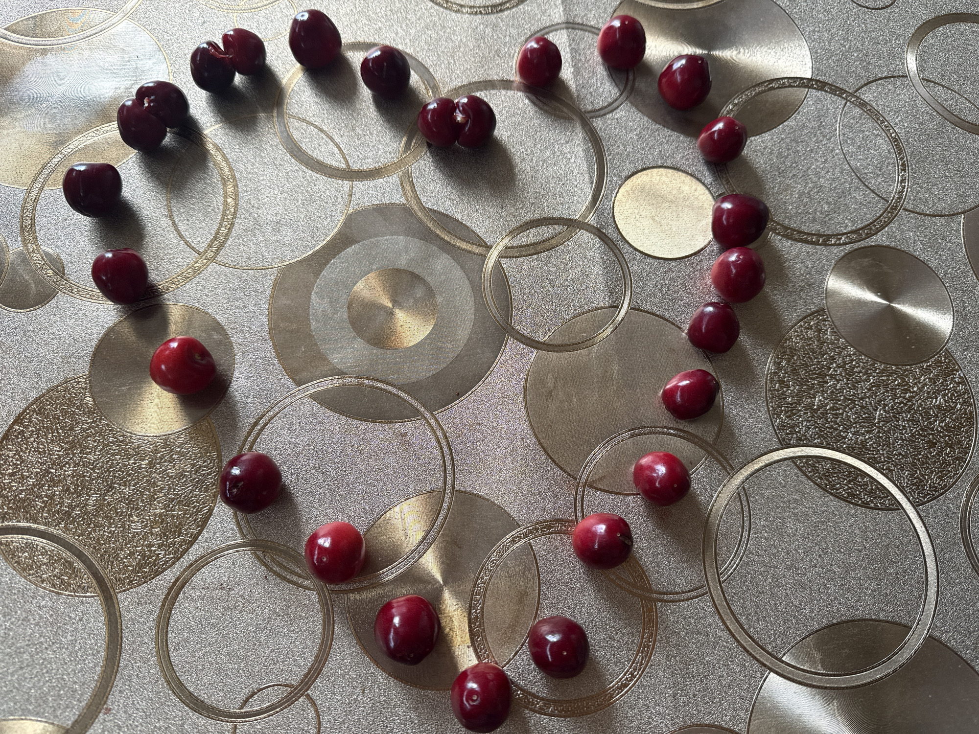 A top-down, medium shot captures a surface adorned with a textured pattern of overlapping circles in gold and silver tones. Scattered across this surface are approximately 30 ripe cherries, primarily clustered towards the center and right side of the frame. The cherries are a deep red hue, with some showing a glossy sheen. Their arrangement forms a rough, incomplete circular or crescent shape. The lighting casts subtle shadows from the cherries onto the surface, indicating a light source from the upper right. There are no people or indications of a specific city or location.