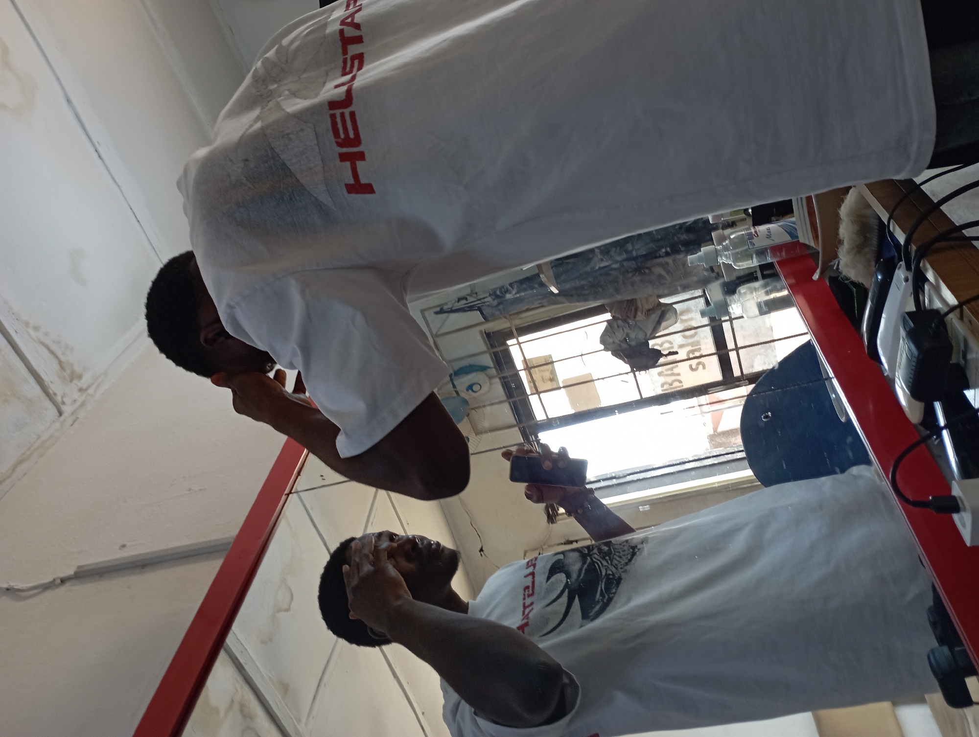 The image, rotated 90 degrees clockwise, captures a scene inside what appears to be a barbershop or salon, primarily through a mirror reflection. A young Black man is depicted, wearing a white t-shirt. On his shirt, the red text "HELLSTAR" is clearly visible, along with a dark graphic design, possibly an animal's head, below it.
In the lower part of the frame (when upright), the man's reflection is seen holding a black smartphone in his right hand, suggesting he is taking a selfie or recording. He looks upward, likely at the phone screen or into the mirror. The upper part of the frame shows the 'real' man from a partial view, looking downwards towards his reflection.
The background visible in the reflection includes a window with light coming through, indicating it is daytime. Reversed text, possibly "SALON" or "BARBERSHOP", is faintly visible through the window, providing a clue about the establishment. On a shelf or counter behind the man, several bottles (likely hair care products) and grooming tools, including what appear to be hair clippers, are arranged. The walls are white, with some visible stains or marks. A prominent red structural element, possibly a metal frame or part of a barber's station, runs along the side and across the middle of the frame.