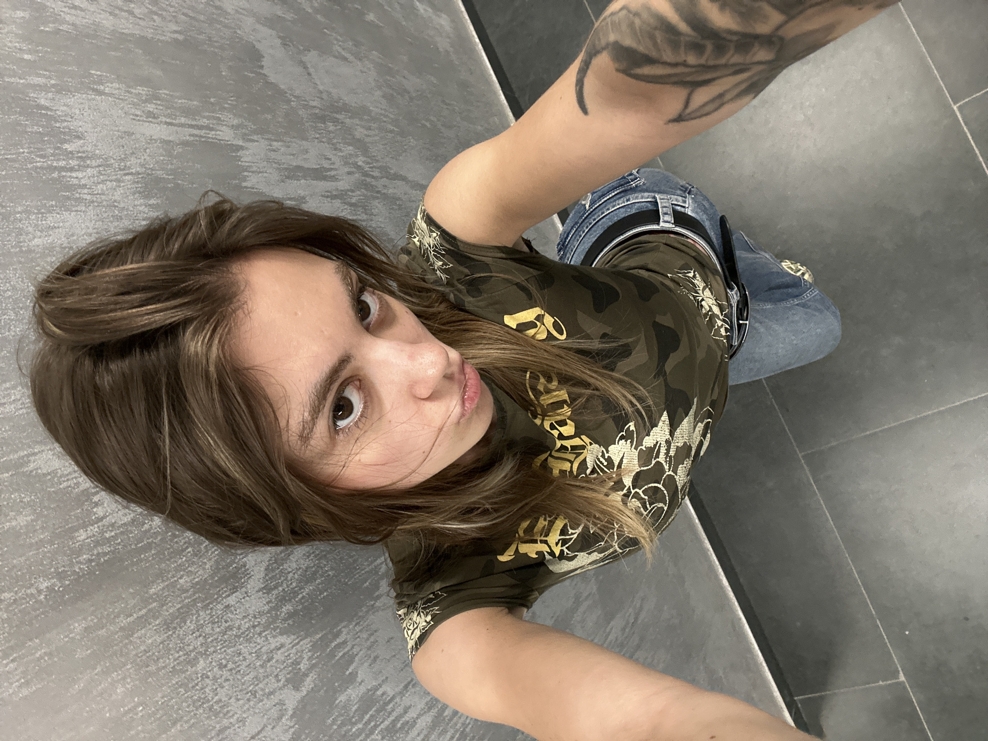A high-angle, medium shot captures a young woman with long, brown hair and brown eyes, looking directly at the camera with a pout. She is wearing a camouflage t-shirt with a gold graphic design and blue jeans with a black belt. Her right arm is extended towards the camera, and a tattoo is visible on her left arm. The background consists of a grey, textured wall and dark grey floor tiles. The setting appears to be indoors, possibly a restroom or changing area, given the utilitarian materials and lighting. There is no indication of the city of Moscow, Russia.