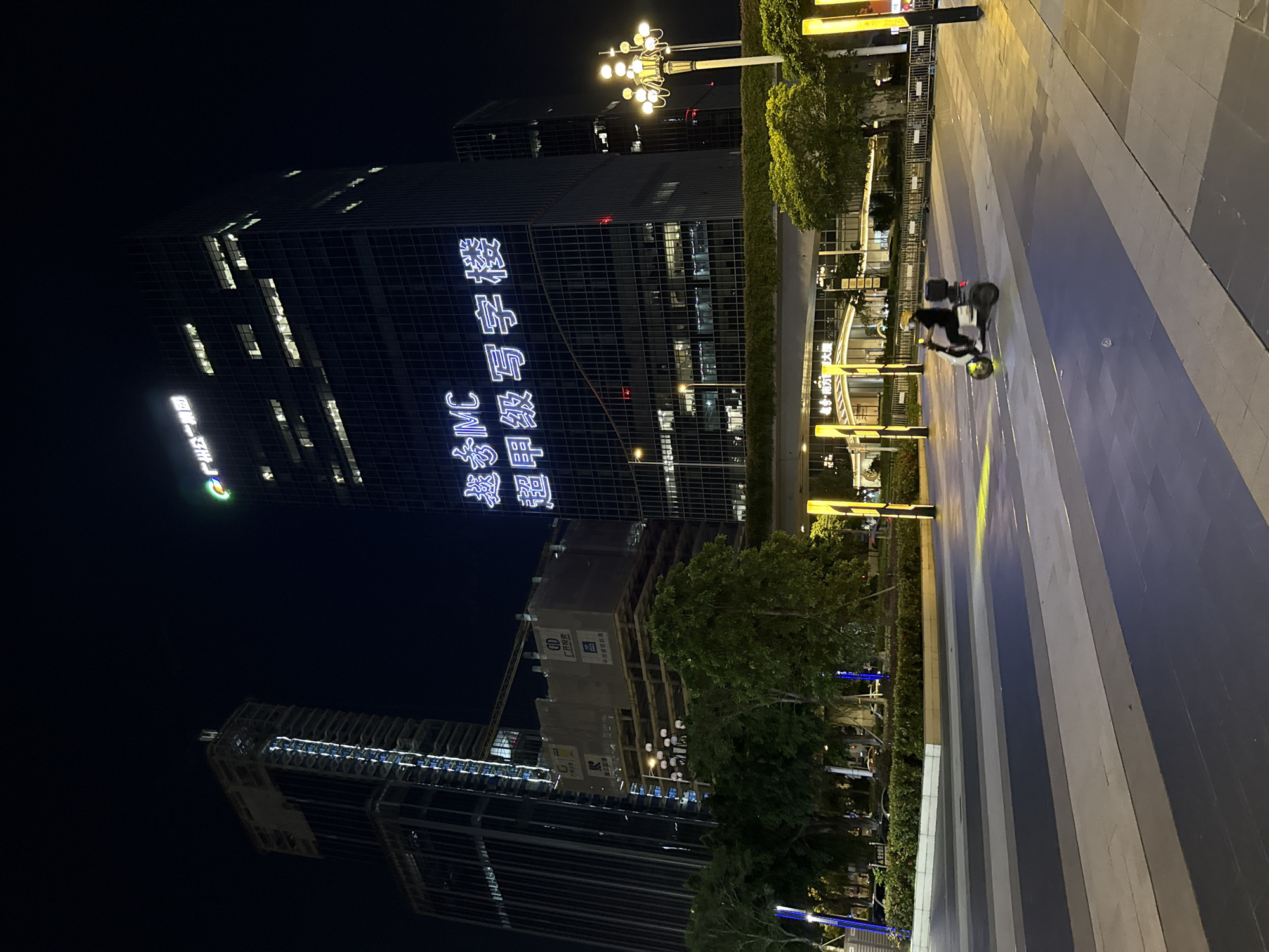 In this nighttime scene in Huangpu District, China, a modern urban environment is depicted. A tall, dark glass skyscraper dominates the left side of the frame, its facade illuminated by white neon lettering that spells out Chinese characters. Below this, a construction site is visible with a crane and scaffolding, indicating ongoing development.
To the right of the buildings, a paved pedestrian walkway features a striped pattern of light and dark gray tiles, reflecting the ambient light. Trees line the walkway, with some areas featuring green hedges. A streetlamp with multiple bulbs stands near the top of the frame, casting a warm glow.
In the middle ground, a person wearing a helmet and dark clothing is riding a scooter with a cargo box on the back. The scooter's wheels are illuminated by a yellow light. The overall atmosphere is one of urban activity at night, with a mix of established infrastructure and new construction. The lighting suggests it is a clear night.