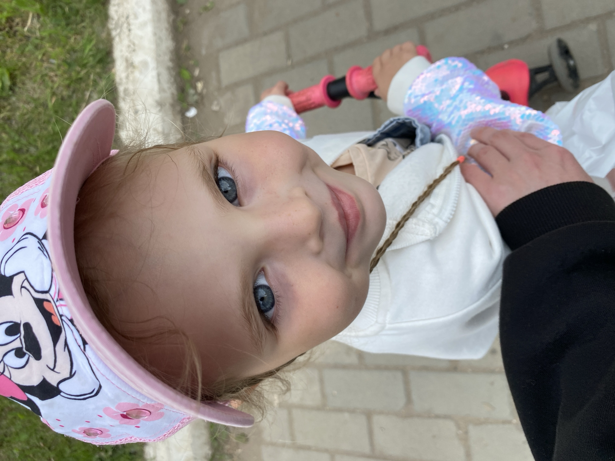 A close-up, high-angle shot shows a young child with striking blue eyes and light brown hair, wearing a white sweater with iridescent sequined cuffs and a pink and white Minnie Mouse baseball cap. The child is holding onto the handlebars of a bright pink scooter. The background consists of light-colored paving stones and green grass, suggesting an outdoor urban environment like a park or sidewalk in Votkinsk, Russia. An adult's arm, wearing black clothing, is visible in the lower left corner of the frame, gently resting on the child. The overall impression is a candid moment of a child enjoying an outdoor activity.