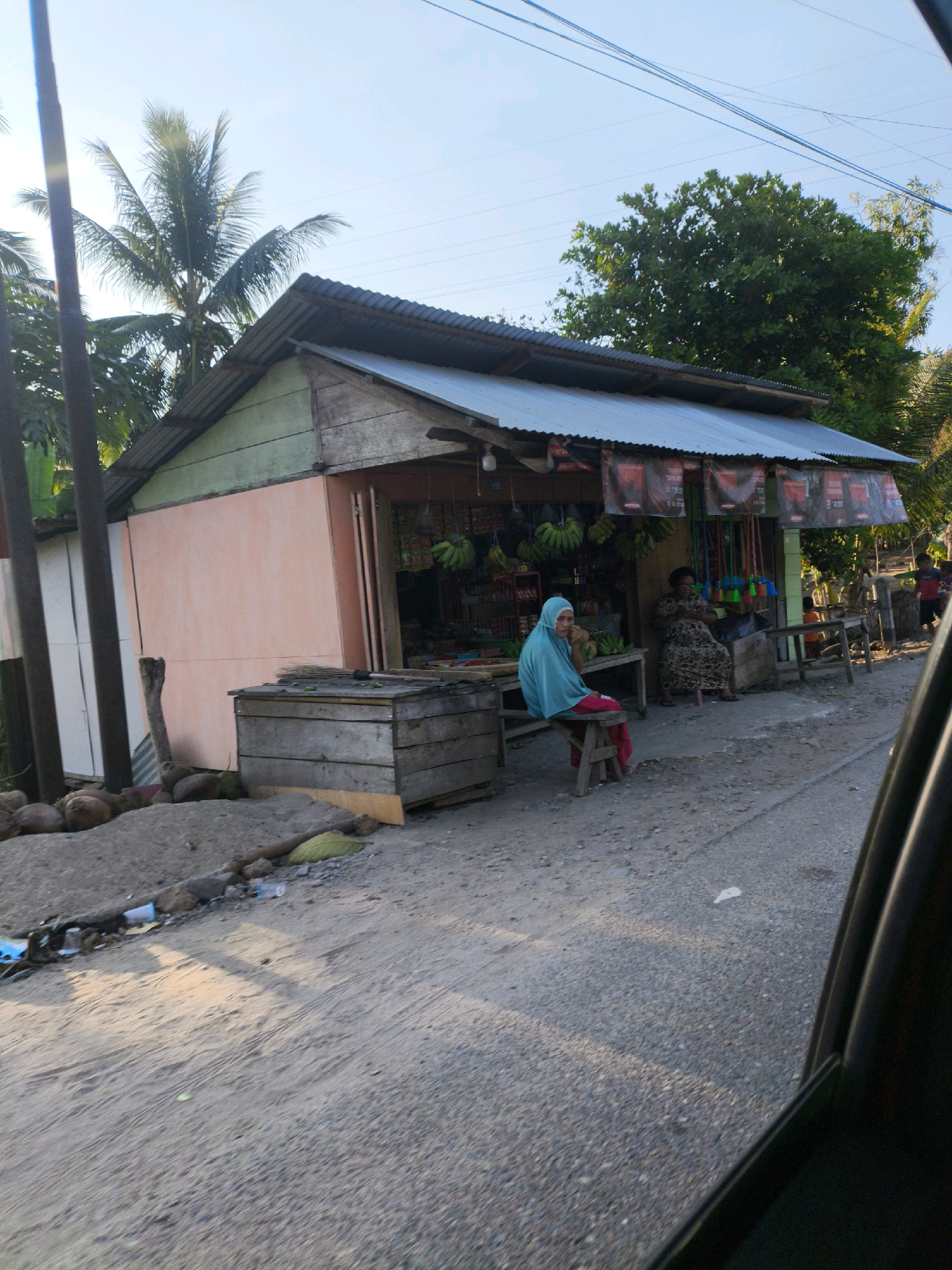 In Dulupi, Indonesia, a small roadside shop stands under a corrugated metal roof. The shop's front is painted a pale peach color, with sections of wood and light green paint visible. A woman wearing a blue hijab and red pants sits on a wooden stool in front of the shop, facing left. Behind her, another woman with dark hair, wearing a patterned dress, is seated indoors. The shop displays various goods, including bunches of bananas hanging from the ceiling. In front of the shop, there are wooden crates, one of which holds brooms and other cleaning supplies. A pile of coconuts is visible to the left of the shop, along with some scattered plastic bottles and debris on the ground. The scene is set on a dusty, unpaved road, with power lines overhead and lush green trees in the background. The lighting suggests it is late afternoon, with sunlight casting long shadows.