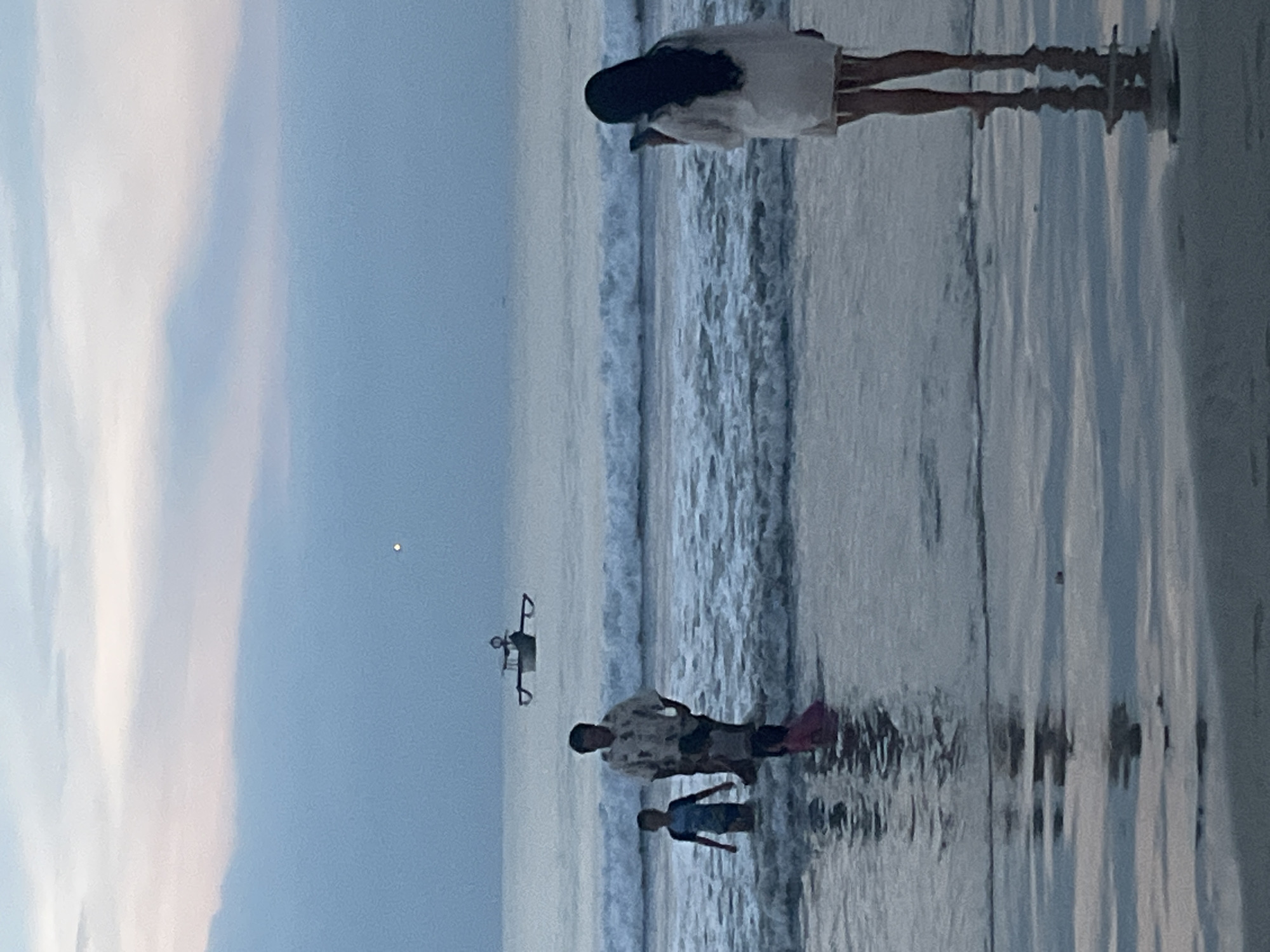The image depicts a twilight scene on Jimbaran Beach, Indonesia. The sky is overcast with a mix of pale blue and faint pink hues, featuring a single bright point of light, likely the moon or a star, visible in the upper sky. The ocean is dark blue-grey with multiple breaking waves producing white foam.
In the mid-ground, a man and two children are in the shallow ocean water, with waves surrounding them. The adult male, wearing a patterned short-sleeved shirt and dark shorts, is positioned towards the center-left, facing slightly towards the shore. To his left, a young child in a dark blue swimsuit is standing waist-deep in the water. A second, younger child, wearing a pink garment, is partially obscured in front of the man, closer to the shoreline.
Further out on the water, beyond the breaking waves, a traditional outrigger boat is visible.
In the foreground, to the right, an adult female with long dark hair stands on the wet sand, facing away from the viewer, towards the ocean. She is wearing a light-colored, possibly white, mid-thigh length cover-up with a fringe hem. She holds a dark-colored smartphone with both hands, appearing to photograph or record the scene before her. Her reflection is visible on the wet, reflective sand.