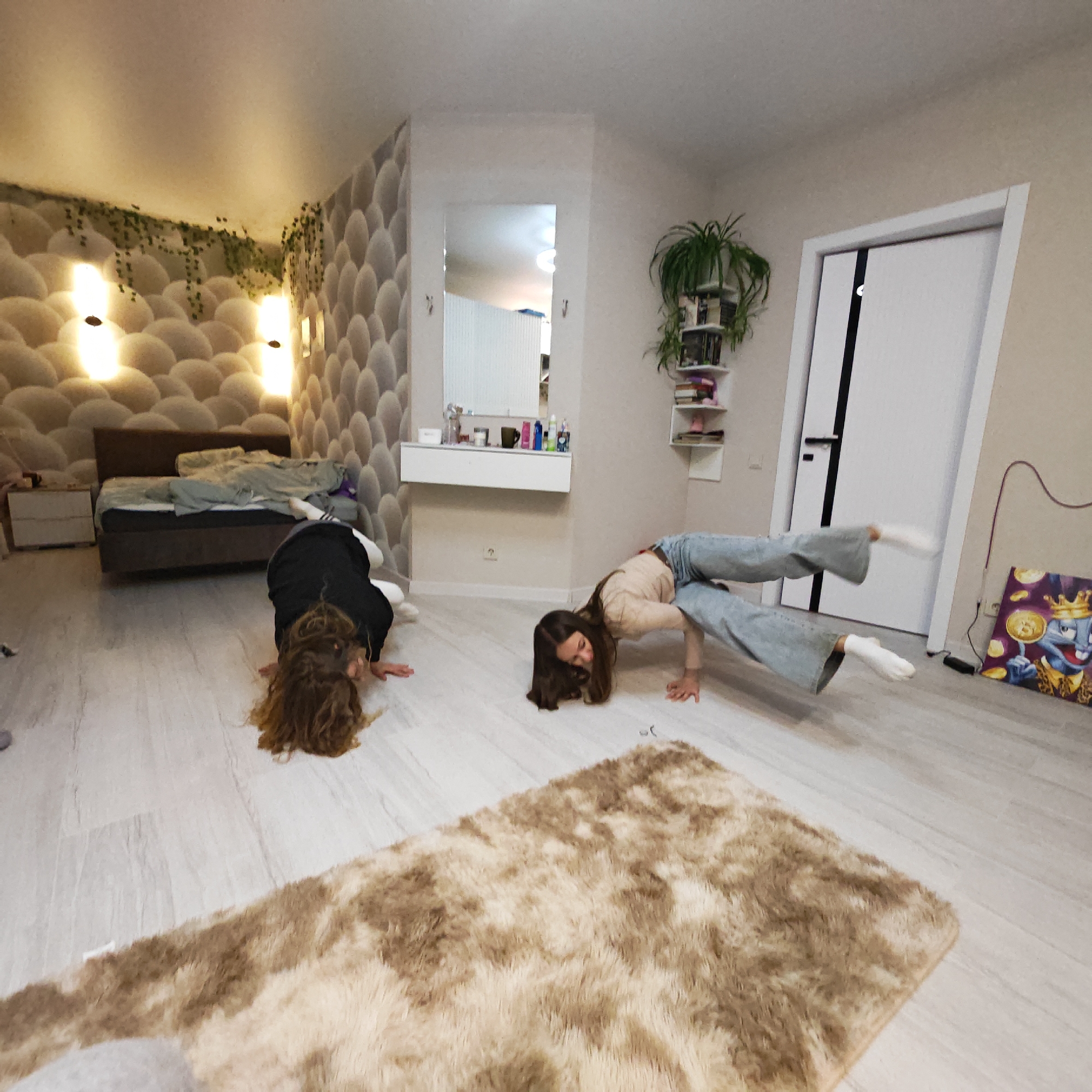 A wide-angle indoor shot depicts two young women performing handstand variations in what appears to be a bedroom setting.
The woman on the right is in a light-colored top and jeans, legs in a split position. The second woman to the left is in a black top and white pants, head resting on the floor.
The room features a bed with a headboard against a textured wall with vertical lights, a vanity with a mirror, and a door. A fluffy rug is in the foreground. An artwork is visible near the door.
There is nothing to indicate the city is Rostov, Russia.