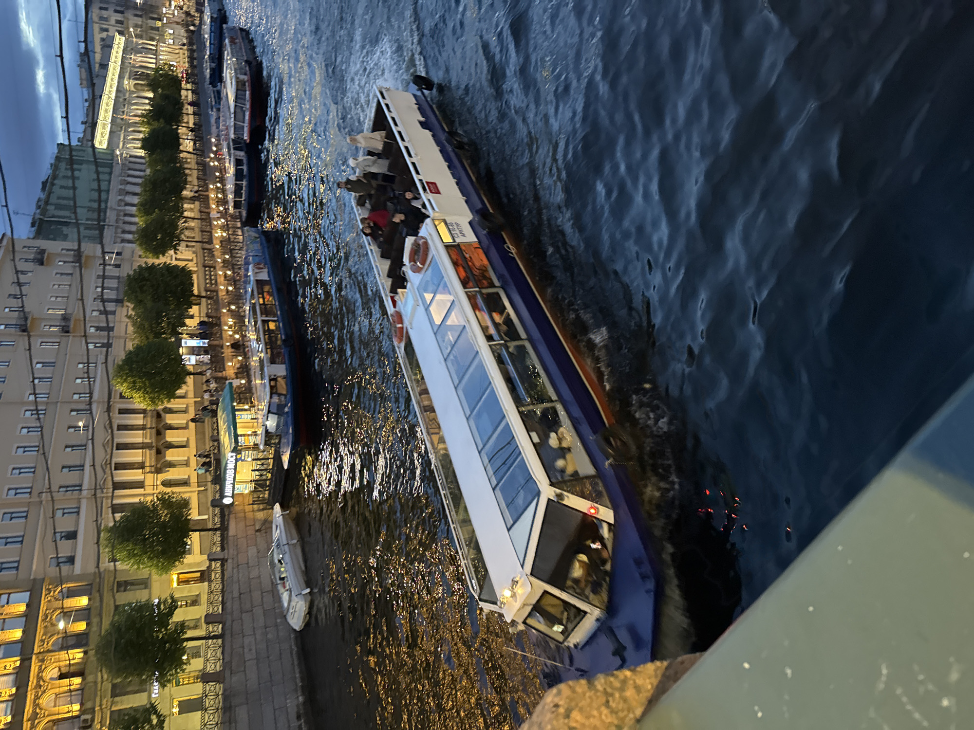 This is a photograph taken at dusk in Saint Petersburg, Russia, likely on the Neva River or one of its canals, given the presence of canal boats and historical architecture. The scene is illuminated by the lights from buildings along the embankment and the glowing water.
In the foreground, a large, blue and white tour boat with a glass roof is navigating the water, creating ripples. The boat is occupied by several passengers visible through the windows and on its open-top deck. On the deck, there are at least five people, some standing and some seated. The passengers are dressed casually.
Further back in the image, several other tour boats are moored or moving along the waterway. These boats are also of a similar design, featuring enclosed cabins and open decks.
The embankment on the left side of the image is lined with historic buildings, some of which are lit up, casting a warm glow. Trees are planted along the embankment between the buildings and the water. The architecture suggests a classical European style, consistent with Saint Petersburg's cityscape. The sky above is dark, indicating late evening or early morning. The water itself is dark and choppy, reflecting the lights from the city.
