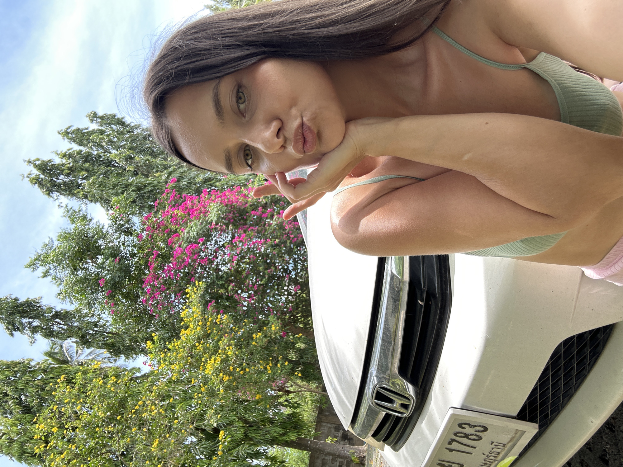 A young woman with long, dark brown hair and green eyes is taking a selfie outdoors. She is wearing a light green ribbed crop top and pink shorts. Her right hand is supporting her chin and her lips are pursed. Behind her, a white Honda car is partially visible. The car has a license plate with Thai characters and the numbers "1783". The background features lush greenery with blooming trees, including yellow and pink flowers, and tall trees. The sky is partly cloudy. The setting appears to be in Baan Mae Nam, Thailand.