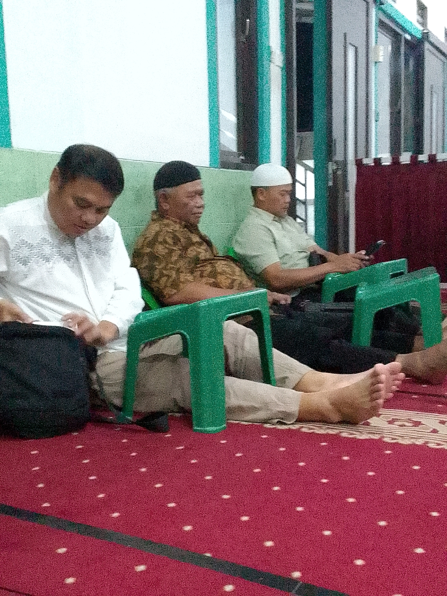 Three men are seated on green plastic chairs on a red carpet with white polka dots. The man on the left wears a white shirt with embroidered details and tan pants. The man in the middle wears a brown patterned shirt and black pants. The man on the right wears a light green shirt, white cap, and black pants, holding a mobile phone in his right hand. The background shows a light green wall with teal stripes, and a dark red curtain. The setting appears to be inside a mosque or similar religious building in Gunungmasigit, Indonesia.