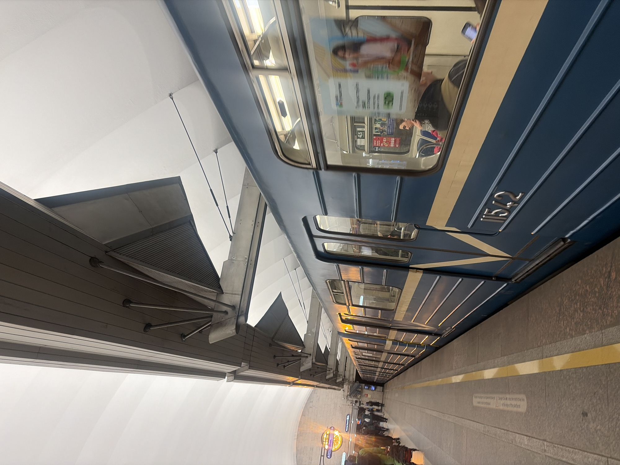 The image captures a modern subway station in Shushary, Russia, with a train stopped at the platform. The perspective is from the platform, looking down the length of the station, with the image rotated 90 degrees clockwise from its natural horizontal orientation.
**Subjects:** The primary subject is a subway train, painted blue with two cream-colored horizontal stripes along its side. The train number "1152" is clearly visible. Reflections in the train's windows show advertisements and the blurry figures of people inside the carriages. On the platform, several people are visible further down, some standing near a circular, brightly lit sign, possibly a station clock or information display.
**Scene/Setting:** The subway station features high, vaulted white ceilings, which contrast with dark grey, possibly metallic, structural elements that support the overhead infrastructure. These structures incorporate slatted designs, suggesting ventilation or lighting elements. The platform floor is made of textured grey paving, marked with a prominent yellow safety line running parallel to the train tracks. The station appears clean, spacious, and brightly lit, indicating either daytime or effective artificial lighting.
**Activity:** The train is stationary at the platform, suggesting passengers are either boarding, alighting, or waiting to do so. People on the platform appear to be waiting for the train or moving along the platform.
**Notable details:** The architecture of the station is contemporary and functional. The overall atmosphere seems orderly.
**Visible text:** On the side of the train, the identification number "1152" is prominent. On the platform wall, a safety warning is displayed in both Russian and English. The English text reads: "Danger! Do not cross the restrictive line on the edge of the platform." The Russian text above it states: "Будьте осторожны и не поддавайтесь давлению толпы. Не пересекайте ограничительную линию на краю платформы." (Translating to: "Be careful and do not yield to the pressure of the crowd. Do not cross the restrictive line at the edge of the platform.")