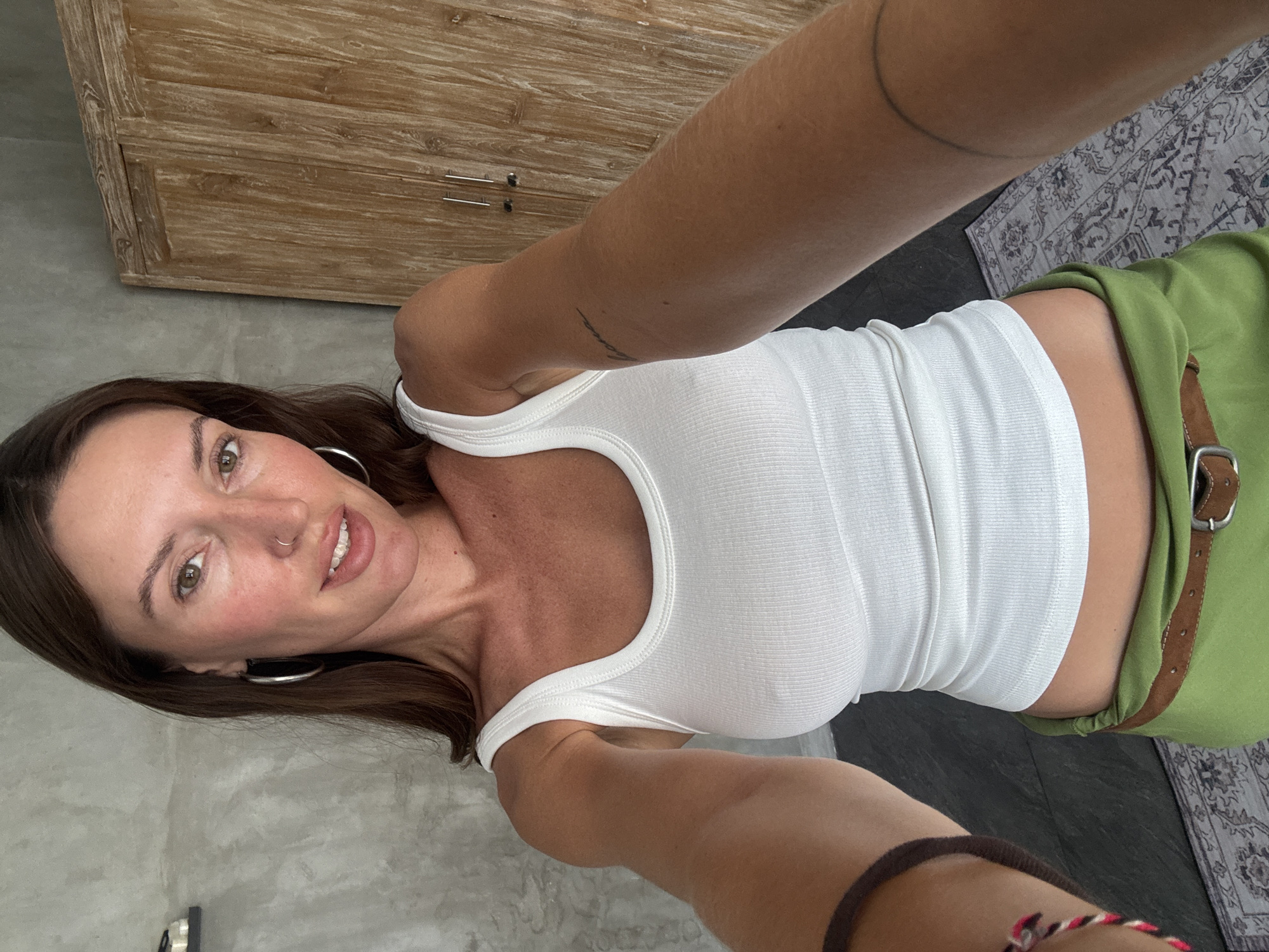 A woman with long brown hair, wearing a white tank top and a green skirt with a brown belt, stands in a room with concrete walls and a wooden cabinet. She is holding up a phone to take a selfie. The room appears to be a bedroom or bathroom. It is a bright, indoor space with natural light. The overall impression is one of a relaxed, personal moment. The location is North Kuta, Indonesia.