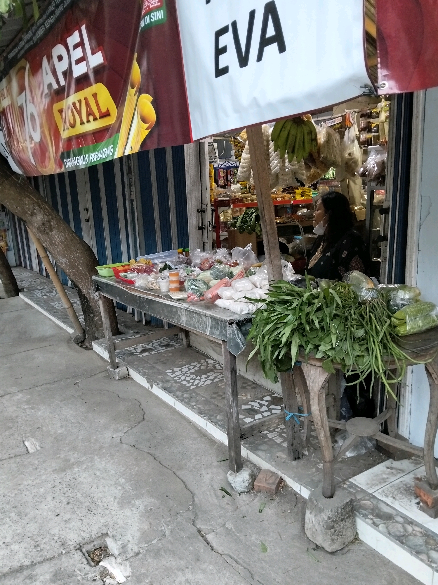 In Kliwonan, Indonesia, a street vendor is observed in front of a shop with a corrugated metal shutter. The vendor, a woman wearing a black garment and a white face mask, sits behind a table laden with various produce and packaged goods. The table itself appears to be constructed from metal, with a wooden surface extending forward and supporting a large bundle of leafy green vegetables and long green beans. Behind the vendor, the shop interior is visible, stocked with shelves of goods. Above the scene, a large banner displays the words "APEL ROYAL" in prominent lettering and the name "EVA." The ground in front of the shop is paved with concrete, showing signs of wear and tear, including cracks and a drainage grate. A large tree trunk is positioned to the left of the vendor's table, leaning against a wooden pole.