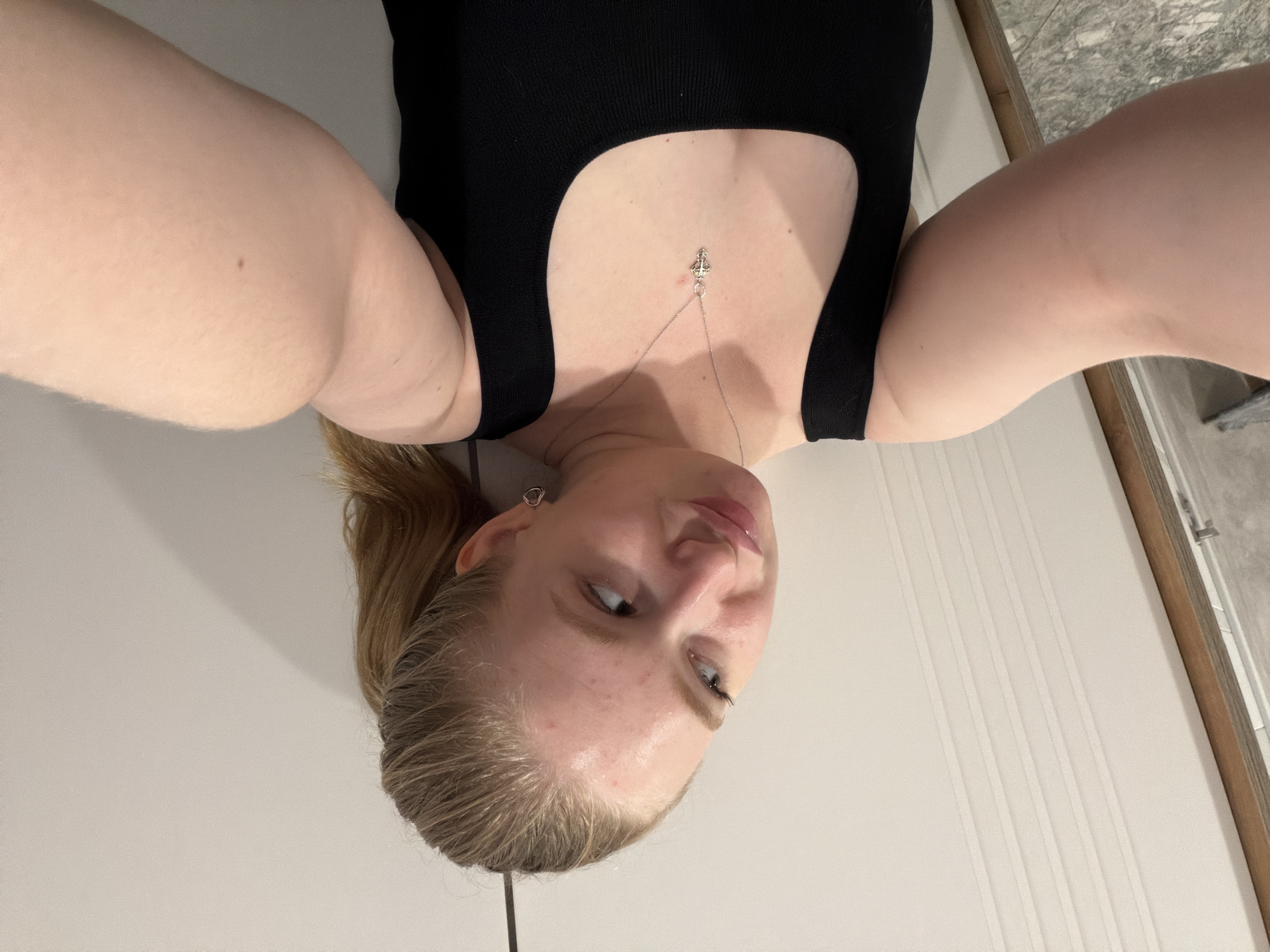 An eye-level, medium selfie shows a young Caucasian woman with blonde hair pulled back in a ponytail and light skin. She is wearing a black tank top and a silver cross necklace. Her head is tilted to the left as she looks off-camera. The background is a plain white wall with vertical grooves. The overall lighting is even and bright, with a slight shadow cast on the wall behind her head. There are no identifiable landmarks or features that would indicate a specific location within Rossiyskiy, Russia.
