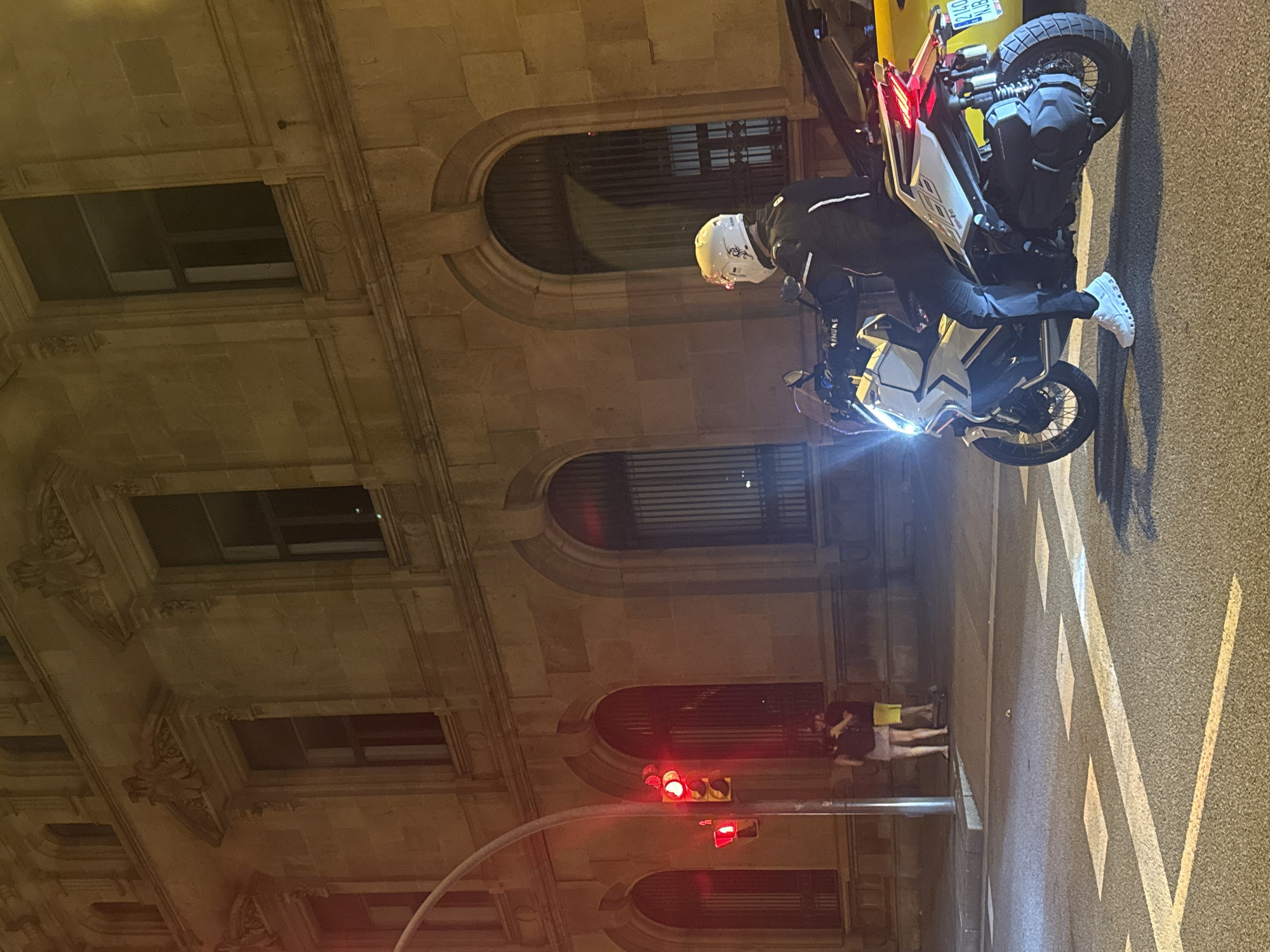 The image depicts a nighttime street scene in Barcelona, Spain, with a focus on a person on a white scooter at a traffic light. The scooter is a modern, sporty model with its headlights and taillights illuminated. The rider is wearing a white helmet and a dark jacket with white accents, along with dark pants and white sneakers. A yellow vehicle is partially visible to the right of the scooter.
In the background, a grand, illuminated building with arched windows and classical architectural details stands imposingly. Two individuals are standing on the sidewalk near a traffic light pole on the left side of the image. The traffic light displays red signals. The street is paved and marked with white lines. The overall atmosphere is urban and nocturnal.
