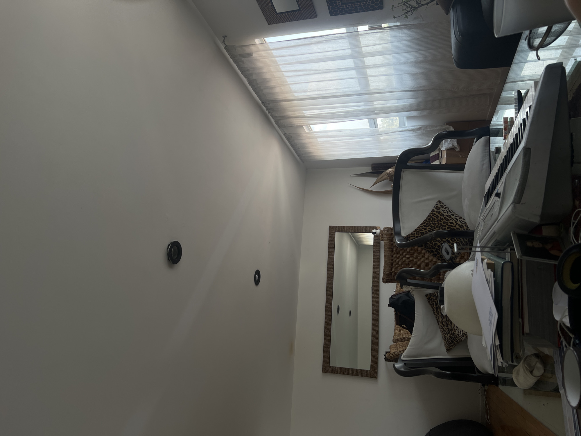 The image shows an interior space in what appears to be a room. The walls are painted white, and two black circular objects, possibly electrical outlets or fixtures, are visible on one wall. A framed mirror hangs on the wall, reflecting what appears to be a narrow passage within the room. To the right, there's furniture, including a chair with a patterned cushion, a keyboard, and other items stacked on a table or shelf. Daylight enters through a window covered by sheer curtains at the top of the image. The angle of the shot indicates that the photo was taken from a low perspective, looking upwards. The location is assumed to be Mexico City, Mexico, based on provided instructions.