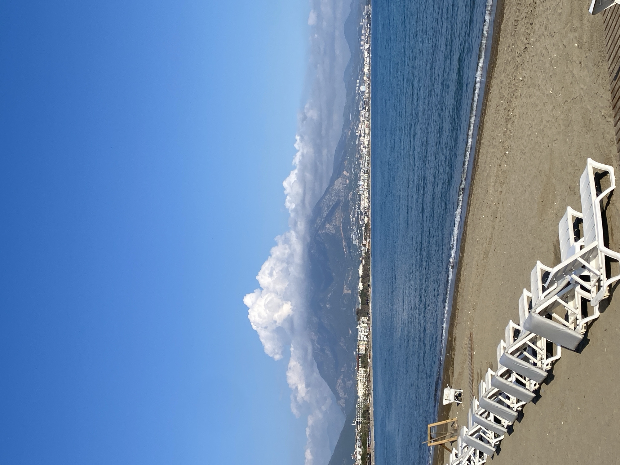 The image, correctly oriented, presents a wide coastal landscape under a clear, bright blue sky. A significant portion of the upper frame is taken up by the uninterrupted sky. In the middle ground, a long mountain range runs parallel to the coast, with some white cumulus clouds clustered around its peaks, particularly on the left side of the frame.
At the base of the mountains, stretching along the coastline, is an urban area composed of numerous buildings, likely hotels, resorts, and residential structures, forming a bustling seaside town. Adjacent to this developed coast lies the calm, blue water of the sea, with gentle waves noticeable as they meet the shore.
The foreground is dominated by a sandy beach. A prominent feature on the beach is a line of white sun loungers or beach chairs, some neatly stacked and others individually arranged, many equipped with grey cushions. These chairs cast distinct shadows, indicating bright sunlight. There are also some wooden structures visible on the sand, possibly related to beach amenities.
The scene depicts a peaceful and sunny day, likely morning or early afternoon given the quality of light and shadows. The weather appears clear, with a few clouds adding texture to the sky over the mountains. No people are visible, and no discernible text is present in the image. The setting is typical of a coastal resort town like Alanya, Turkey.