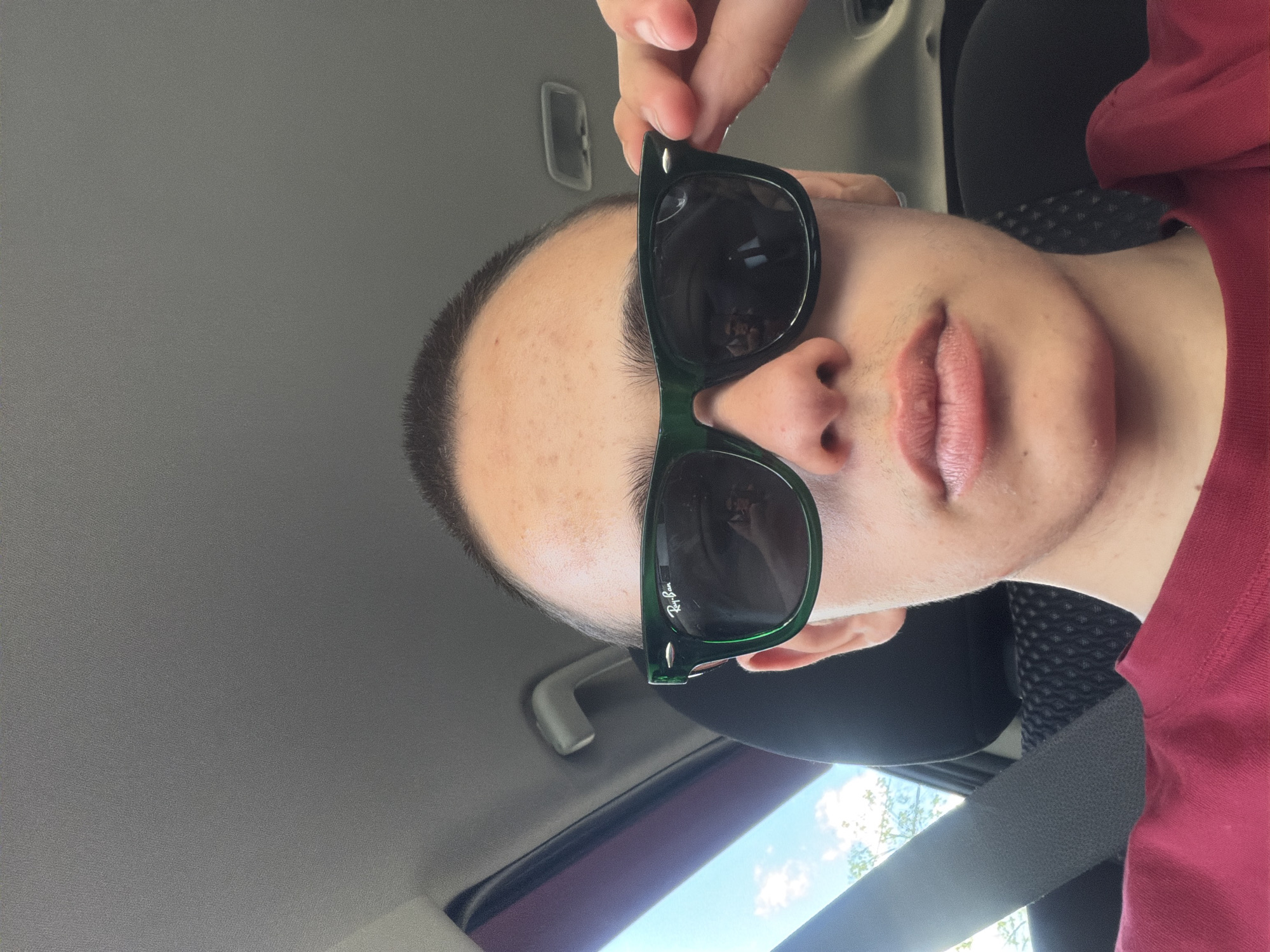 A young person with short hair and sunglasses is in the driver's seat of a car, looking at the camera. They are wearing a red shirt and have their hand on the side of their sunglasses. The car interior is visible, with a seatbelt and part of the dashboard. Outside the car, the sky is visible with some clouds.