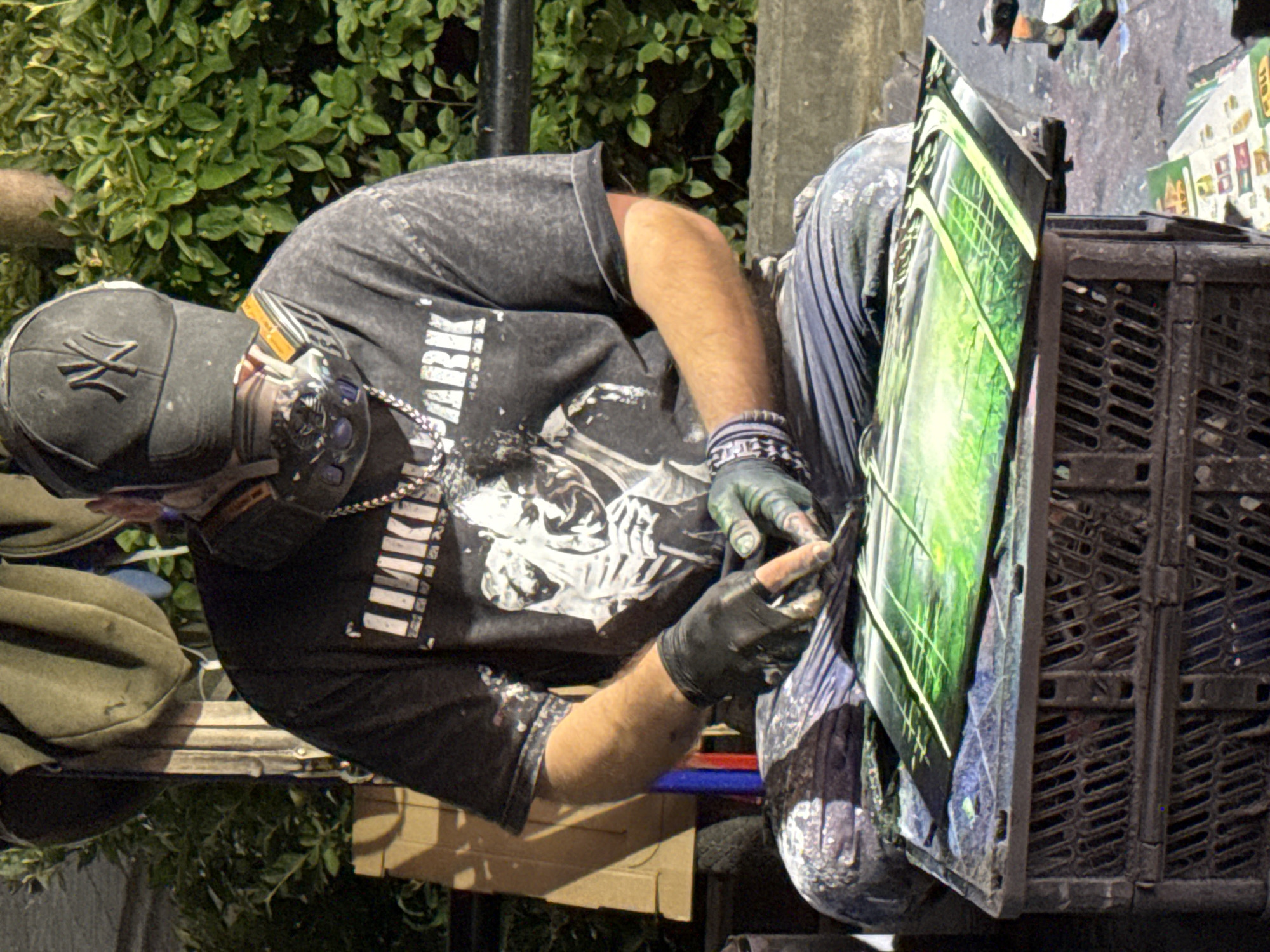 A person, wearing a black baseball cap with a white logo, a black t-shirt with a white graphic print and text, a respirator mask, black gloves, and blue jeans, is sitting down and appears to be working on a painting. The person is surrounded by various items, including what looks like a black plastic crate, some cardboard, and what could be spray cans or art supplies. The background consists of greenery and what appears to be a fence or railing. The lighting suggests it is either evening or a dimly lit indoor space. No specific location in Kazan, Russia, can be identified from the provided image.