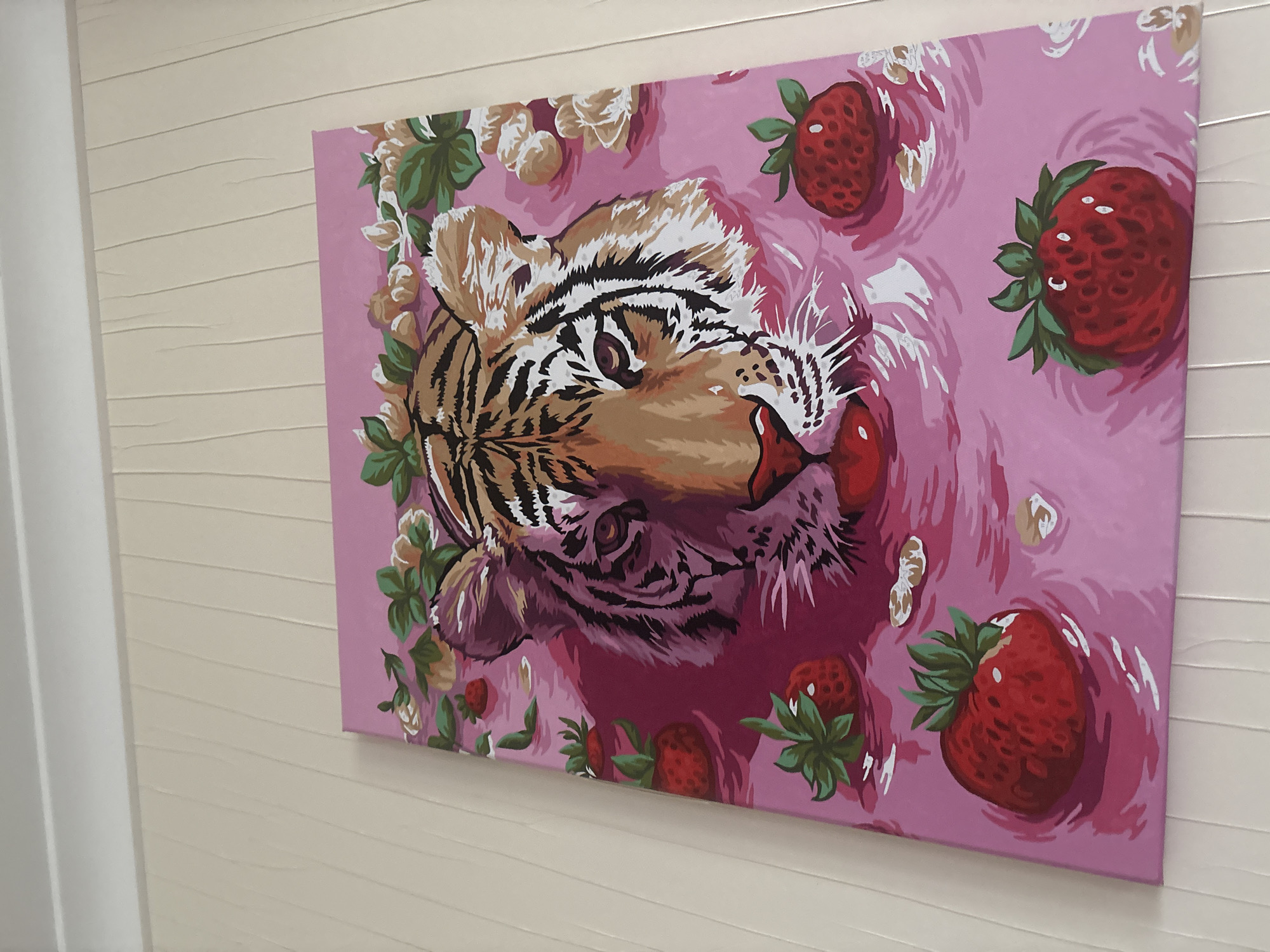 A large canvas painting, positioned horizontally, is prominently displayed on a light beige, horizontally textured wall. The painting features a stylized depiction of a tiger's head partially submerged in a vibrant pink liquid, possibly water or a fruit-infused bath. The tiger's head is central, with its eyes and snout visible, rendered in shades of brown, orange, black, and white. Its mouth is slightly open, revealing teeth.
Scattered around the tiger in the pink liquid are several bright red strawberries with green leaves. There are also floral elements, including leaves and stylized flowers in shades of green, cream, and brown, framing parts of the tiger's head and floating in the liquid. The painting has a graphic, illustrative style, characterized by distinct color blocks and outlines, reminiscent of a paint-by-numbers artwork.
To the left of the painting, a vertical white architectural element, likely a door frame or wall corner, is visible. The room appears to be well-lit by ambient indoor lighting. There is no visible text in the image. The location context is Ореховка, Russia.
