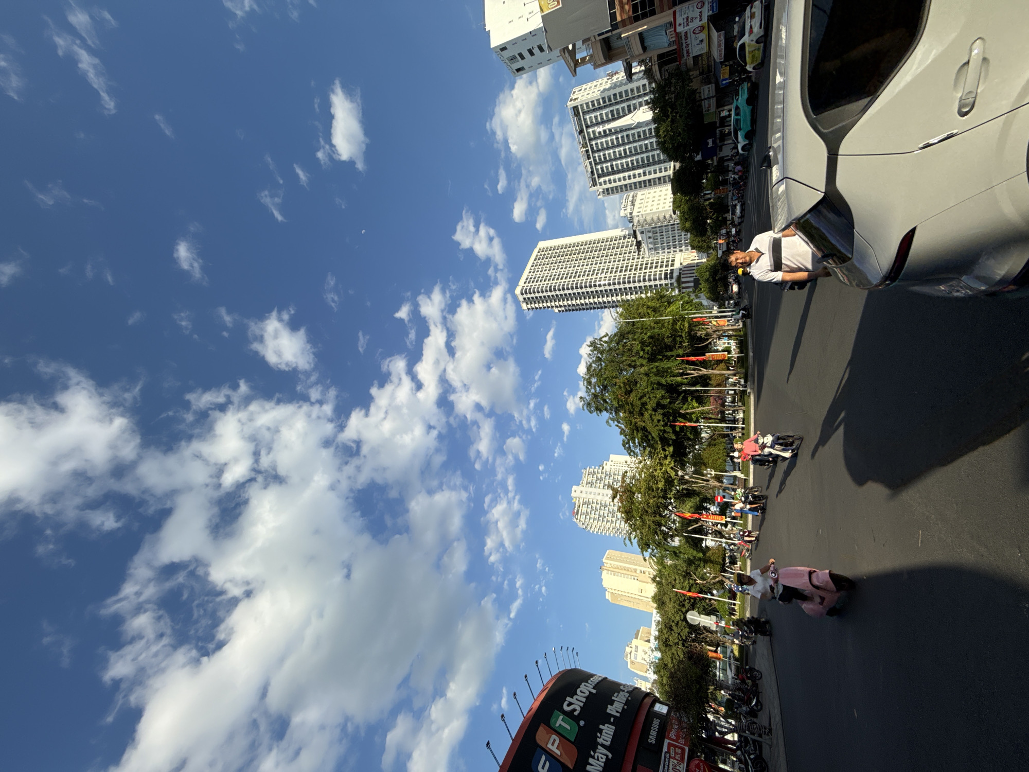 This is a street-level, wide-angle photograph taken in Nha Trang, Vietnam, likely during the daytime given the clear blue sky with scattered white clouds and the shadows cast on the road. The scene depicts a busy urban environment with several tall buildings in the background, including white, multi-story apartment or office buildings and a curved, red and black billboard on the left displaying text in Vietnamese and English, including "FPT Shop". The foreground shows a paved road with several people on motorcycles and scooters. A man in a striped polo shirt is standing next to a light-colored car on the right side of the road. Trees line the street, and there are several red and yellow flags visible. The overall impression is of a sunny day in a commercial district of the city.