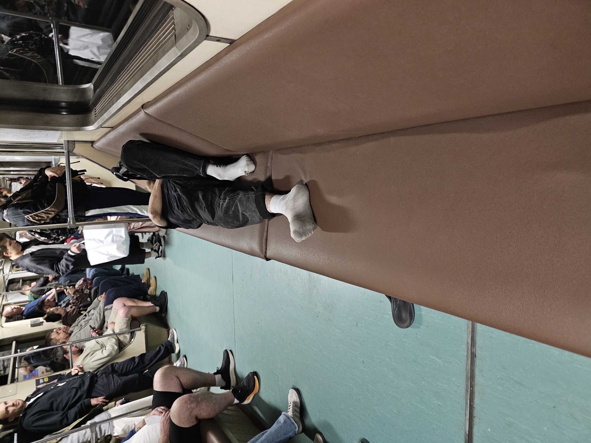 The image depicts the interior of a subway car with several passengers. The perspective is from inside the car, looking down the aisle. On the right side, a person is lying down across the seats, with their legs extended. They are wearing dark pants and white socks, with one sock appearing to have a hole. The seats are upholstered in a brown material. On the left side of the car, several passengers are seated or standing. One man in the foreground on the left is wearing black shorts and black athletic shoes. Other passengers are visible further down the car, some holding bags. The floor of the subway car is a light blue-green color. The overall impression is of a crowded public transportation setting.